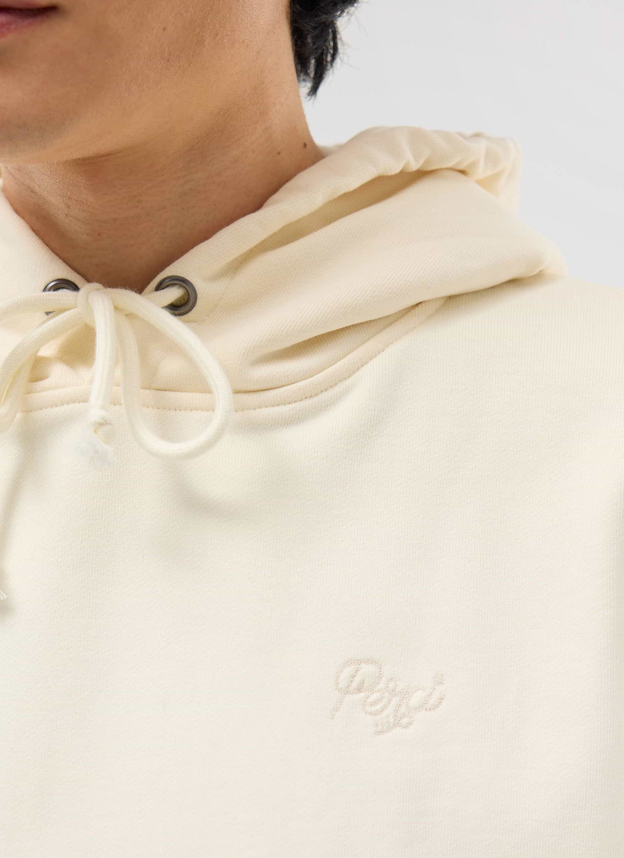 A close-up of a model wearing the Percival Perci Chainstitch Hoodie in ecru, showing the tonal chest embroidery.