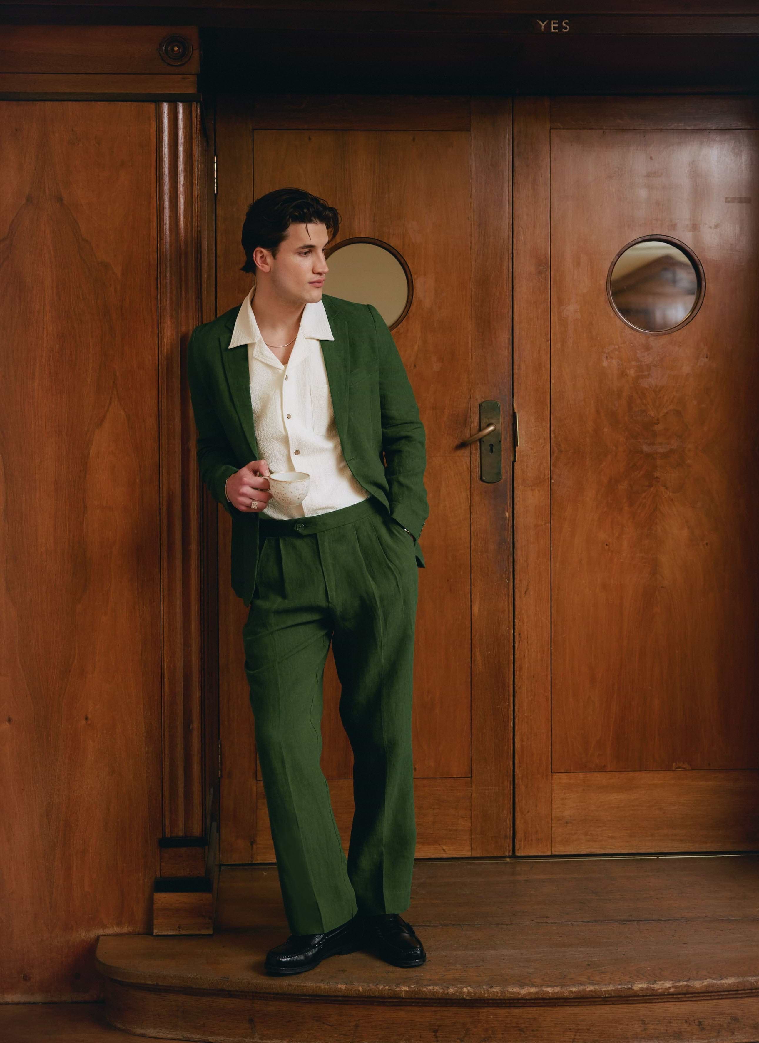 A model wears the Percival Tailored Linen Blazer and matching trousers in forest green, leaning against wooden doors.