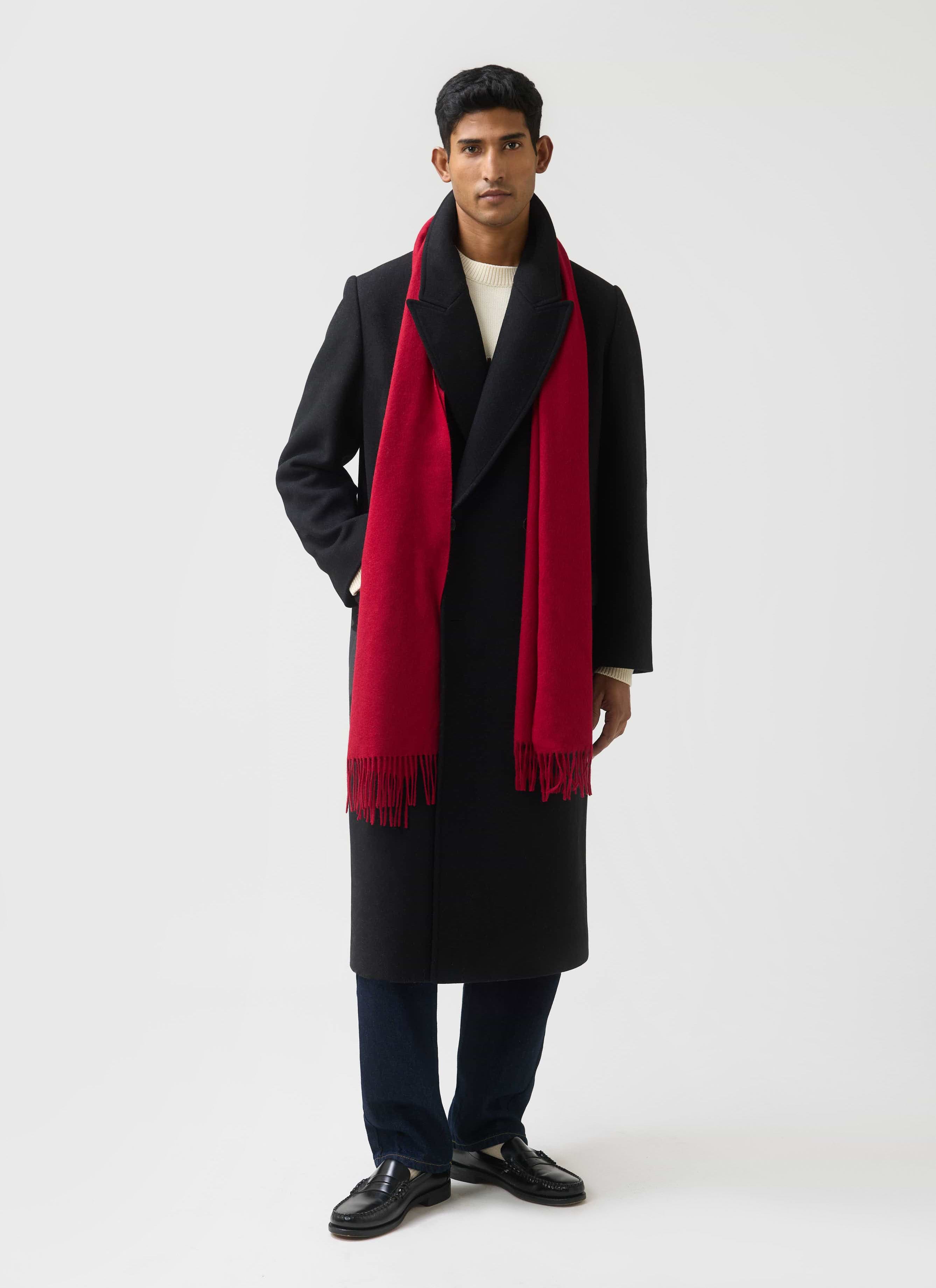 A man models the red Tolkein wool scarf from Percival, styled with a long black overcoat and a cream jumper.