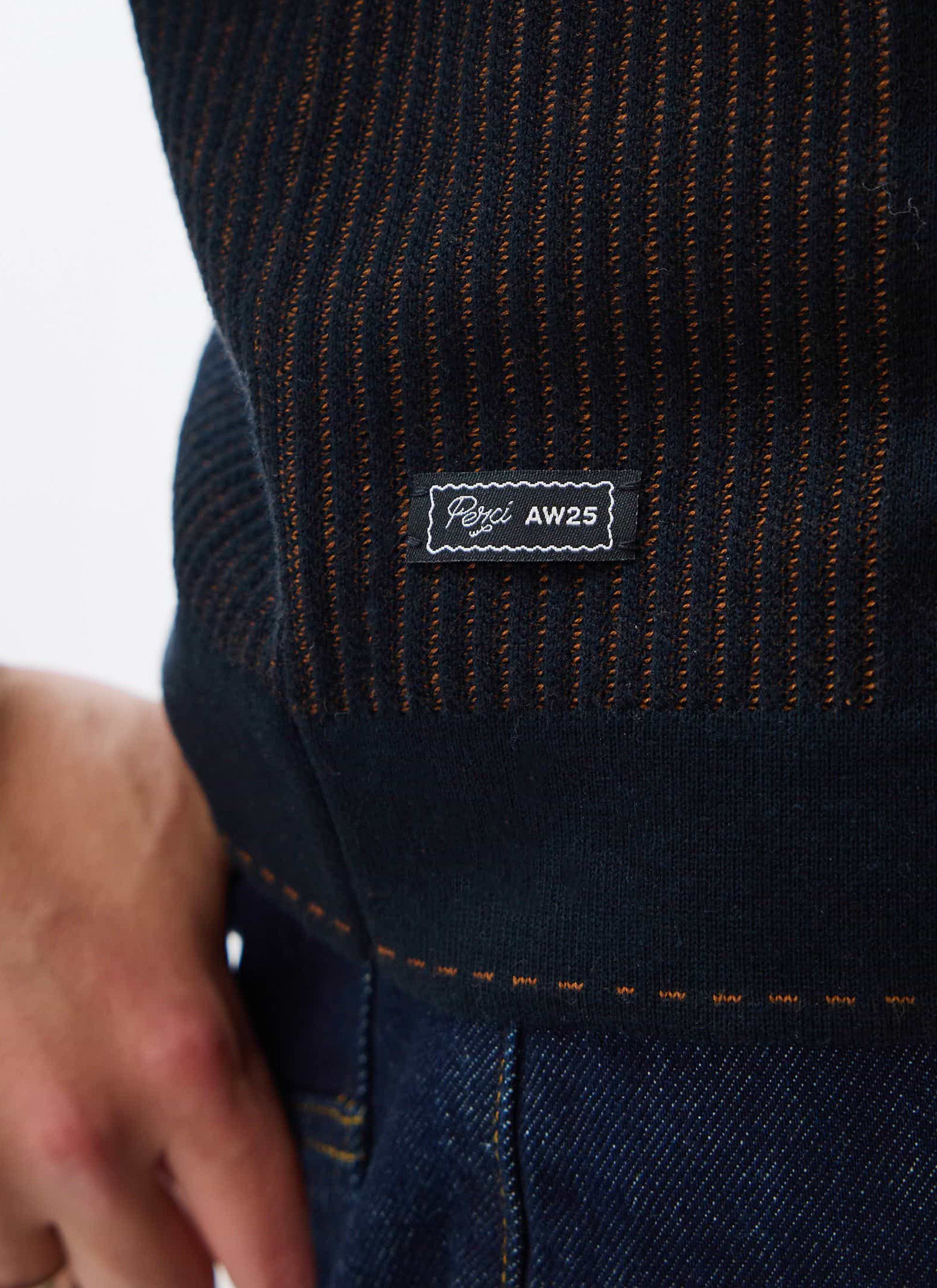 A close-up of the black and brown jacquard knit on the Percival Appendix shirt, showing the branded AW25 label detail.