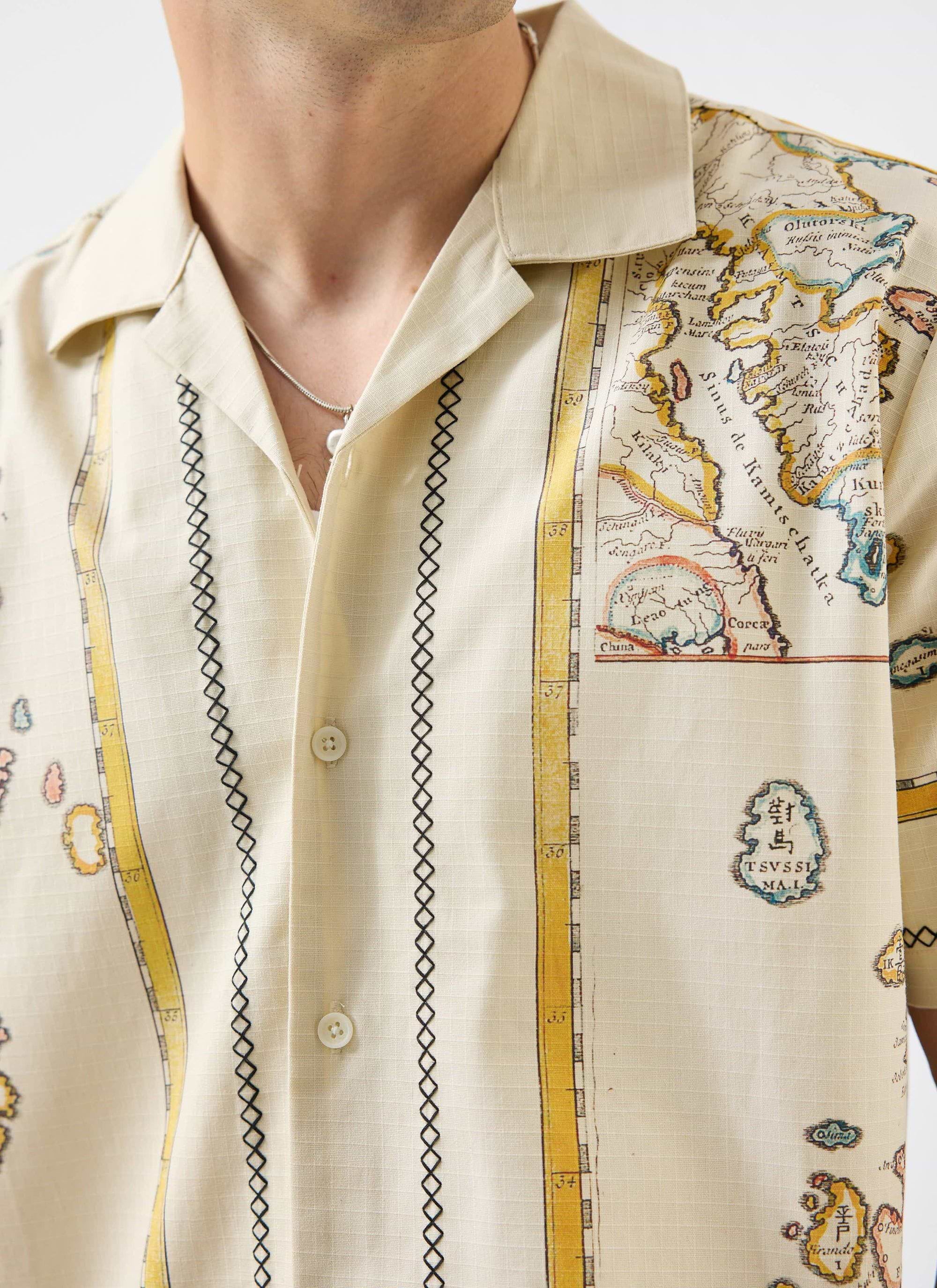 A close-up shot of a model wearing the Percival Ex Libris Cuban Shirt, showing the ecru map print and embroidery details.