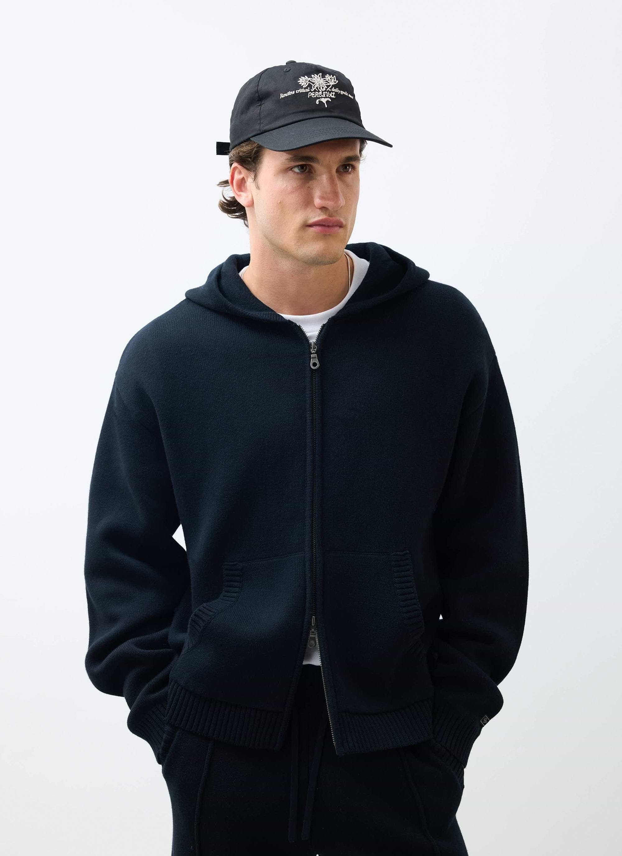 A man models the black Percival Chronicle Knitted Hoodie, a cotton zip-up style, worn with a cap in a studio.