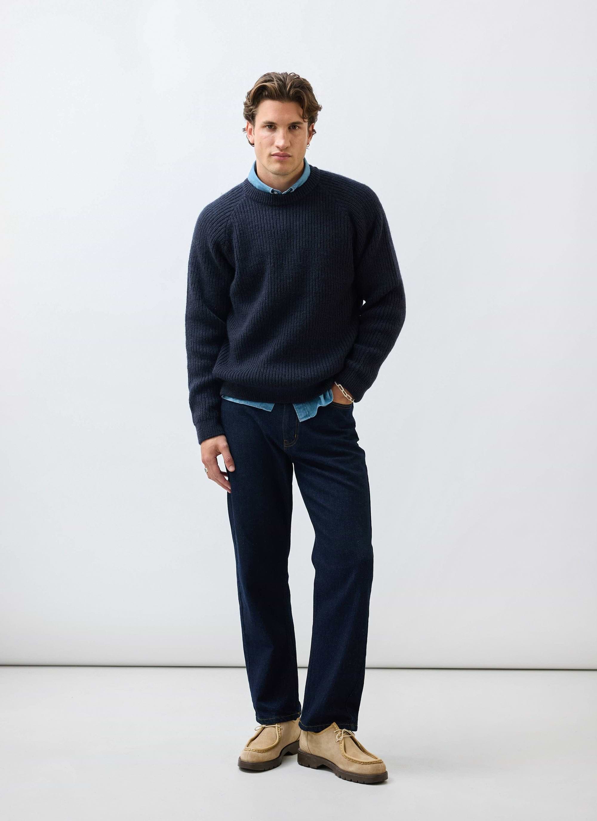 A model styles the Percival Fisherman Raglan Jumper in navy wool over a blue shirt with dark denim jeans.