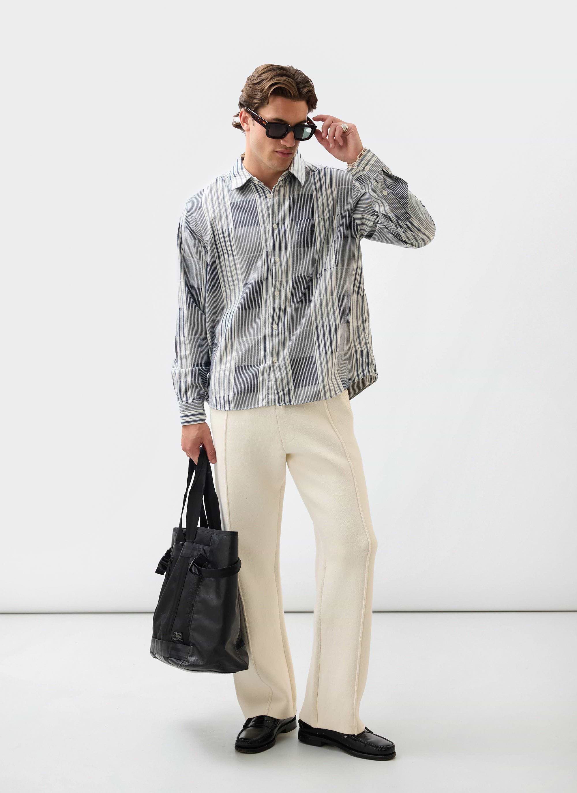 A model wears the Percival Chronicle Knitted Trackpant in ecru, styled with a striped shirt and loafers.