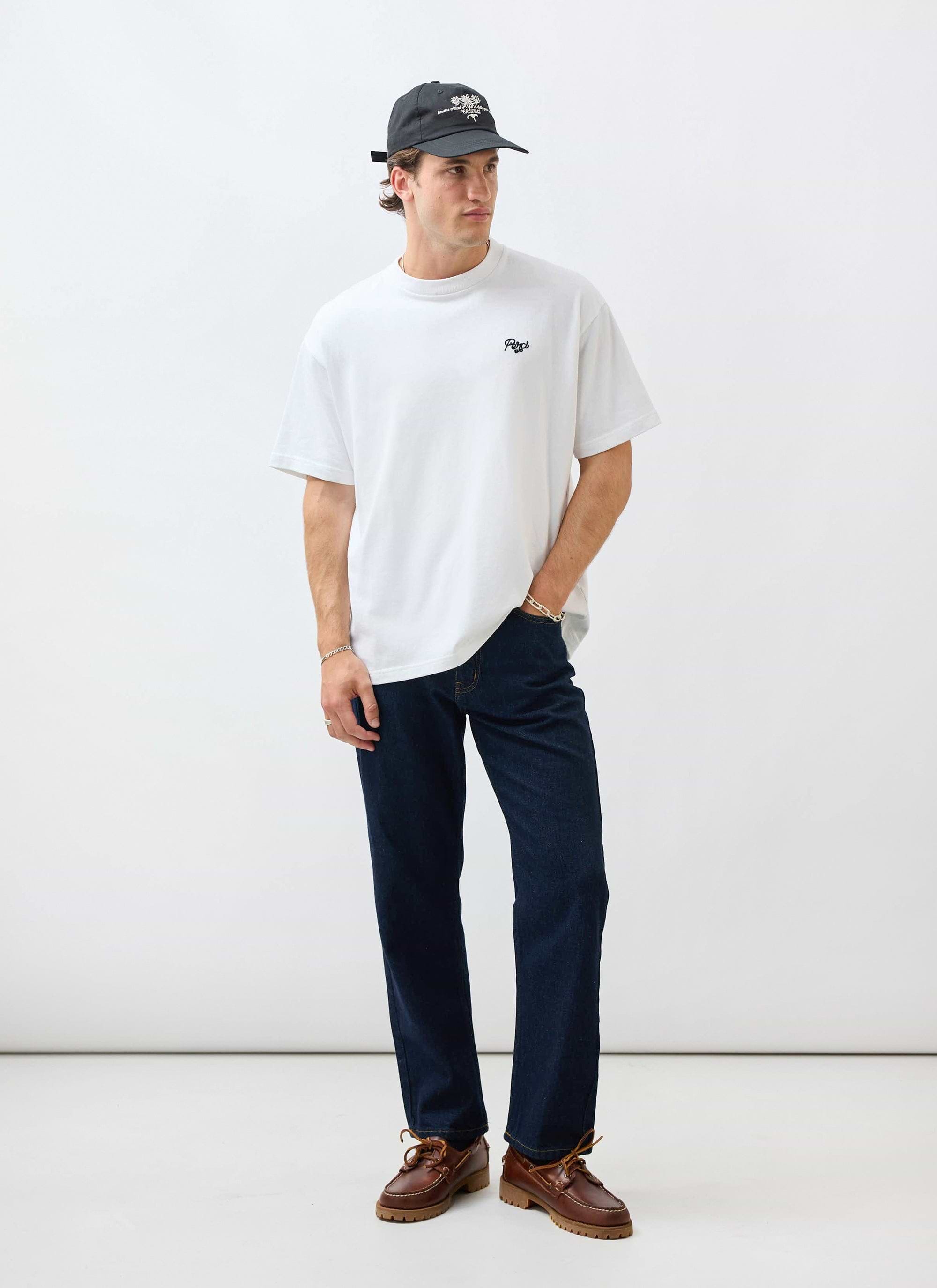 A model wears the Percival Du Temps Oversized T Shirt in white, styled with dark wash jeans, brown shoes and a black cap.