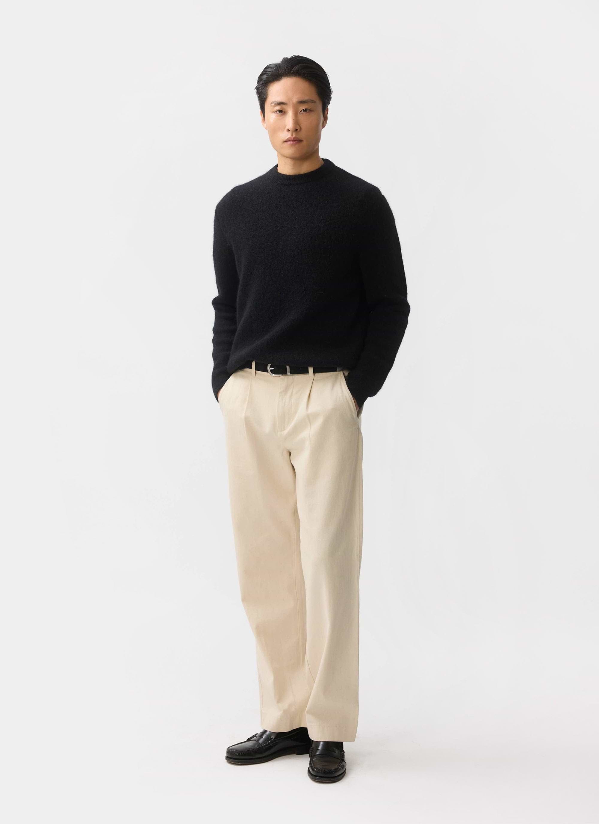 A man models the black mohair White Glove Crew Neck Jumper by Percival with cream trousers against a white background.