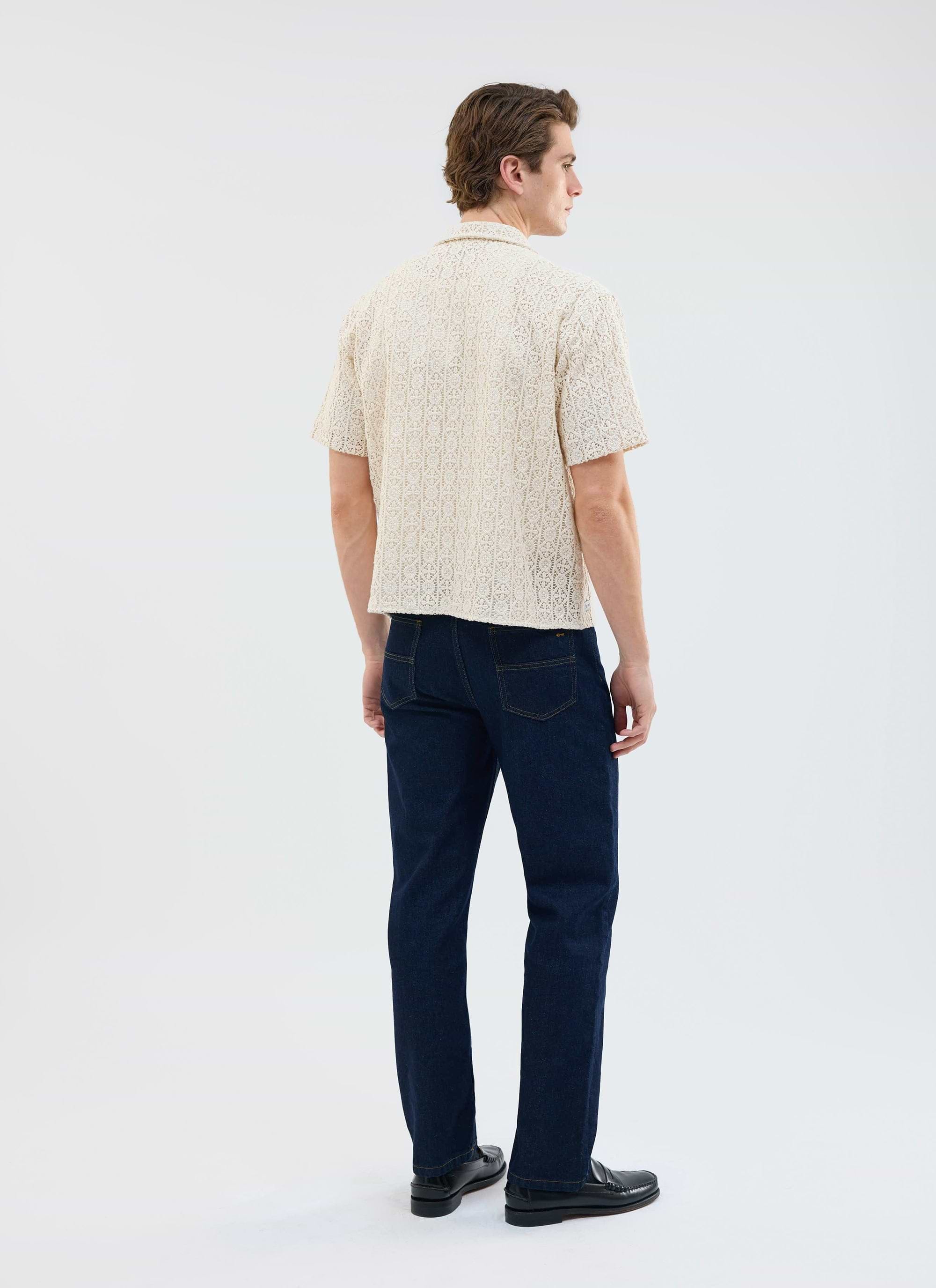 A model, seen from behind, wears the Percival Elio Crochet Cuban Shirt in ecru with dark wash jeans and black loafers.