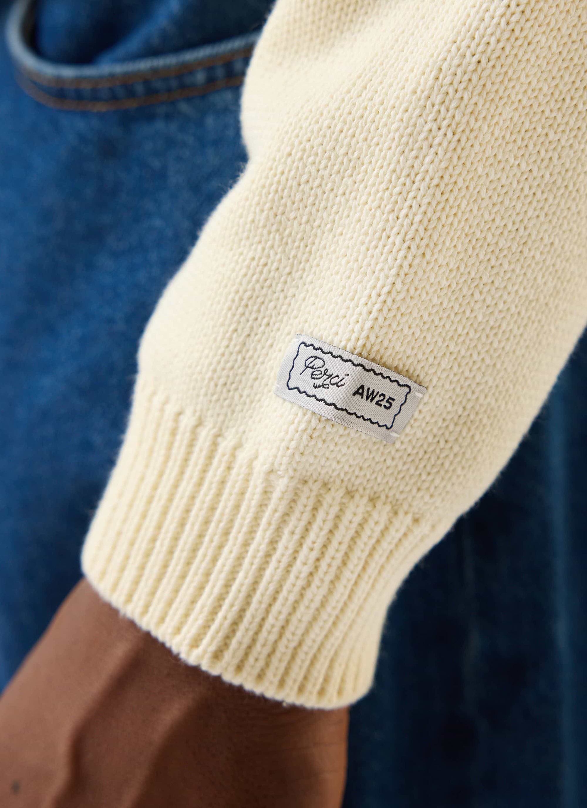 Close-up of the Percival Public Library Knitted Jumper in ecru cream, showing the ribbed cuff detail and branded label.