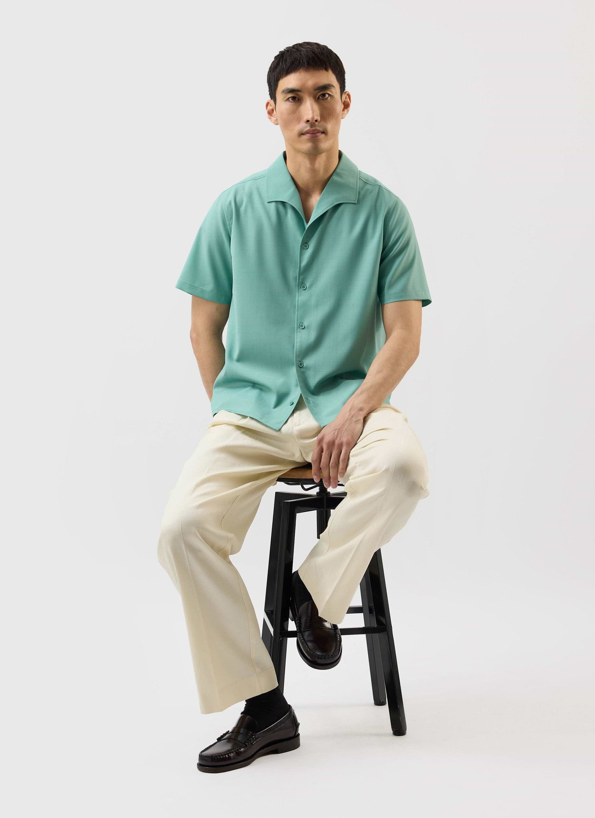 A model sits on a stool wearing the Percival x Warren Drape Cuban Shirt in mint green with cream trousers.