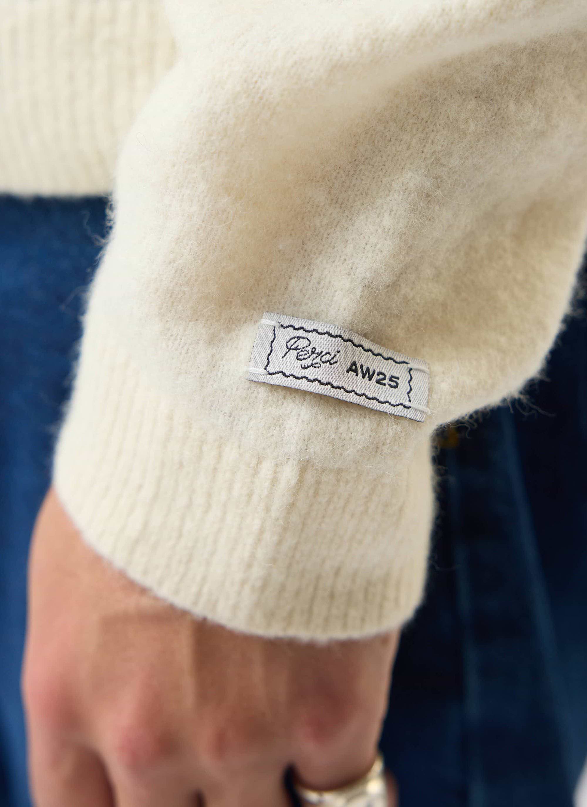 Close-up of the Percival branded tab on the cuff of the ecru Archive Wool Knitted Collared Cardigan.