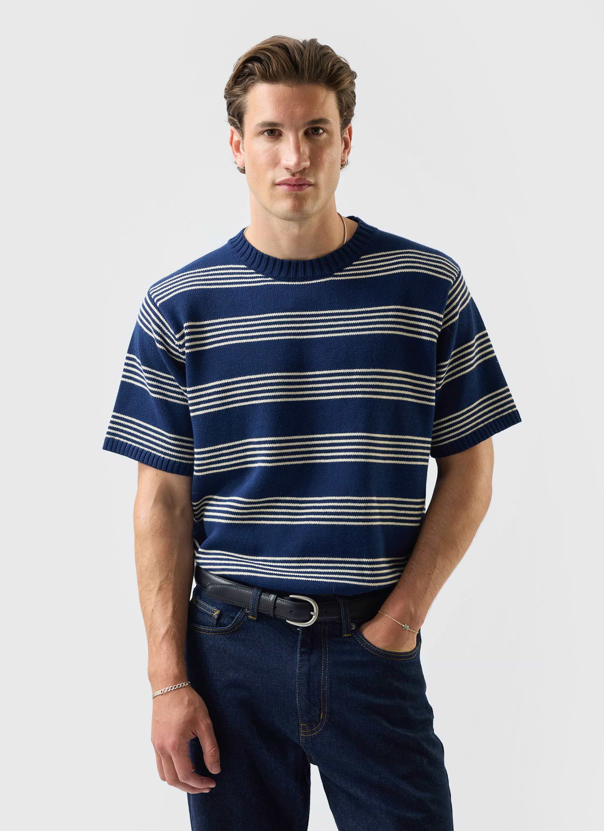 A model wears the Percival Fiction knitted t-shirt in navy with cream stripes, paired with dark denim against a white wall.