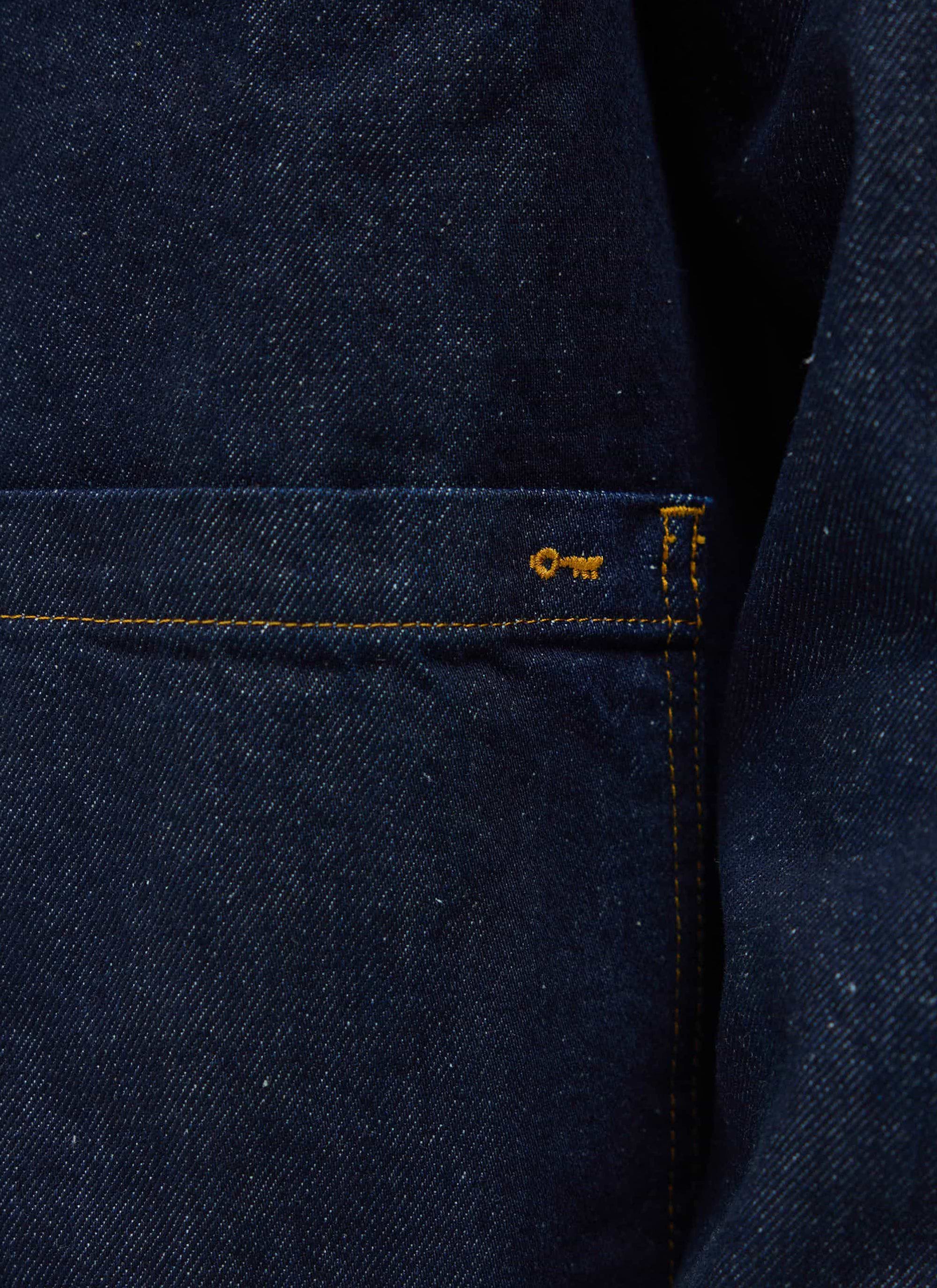 A close-up of the Percival Gove Chore Jacket in indigo denim, showing the patch pocket with contrast stitching and key logo.