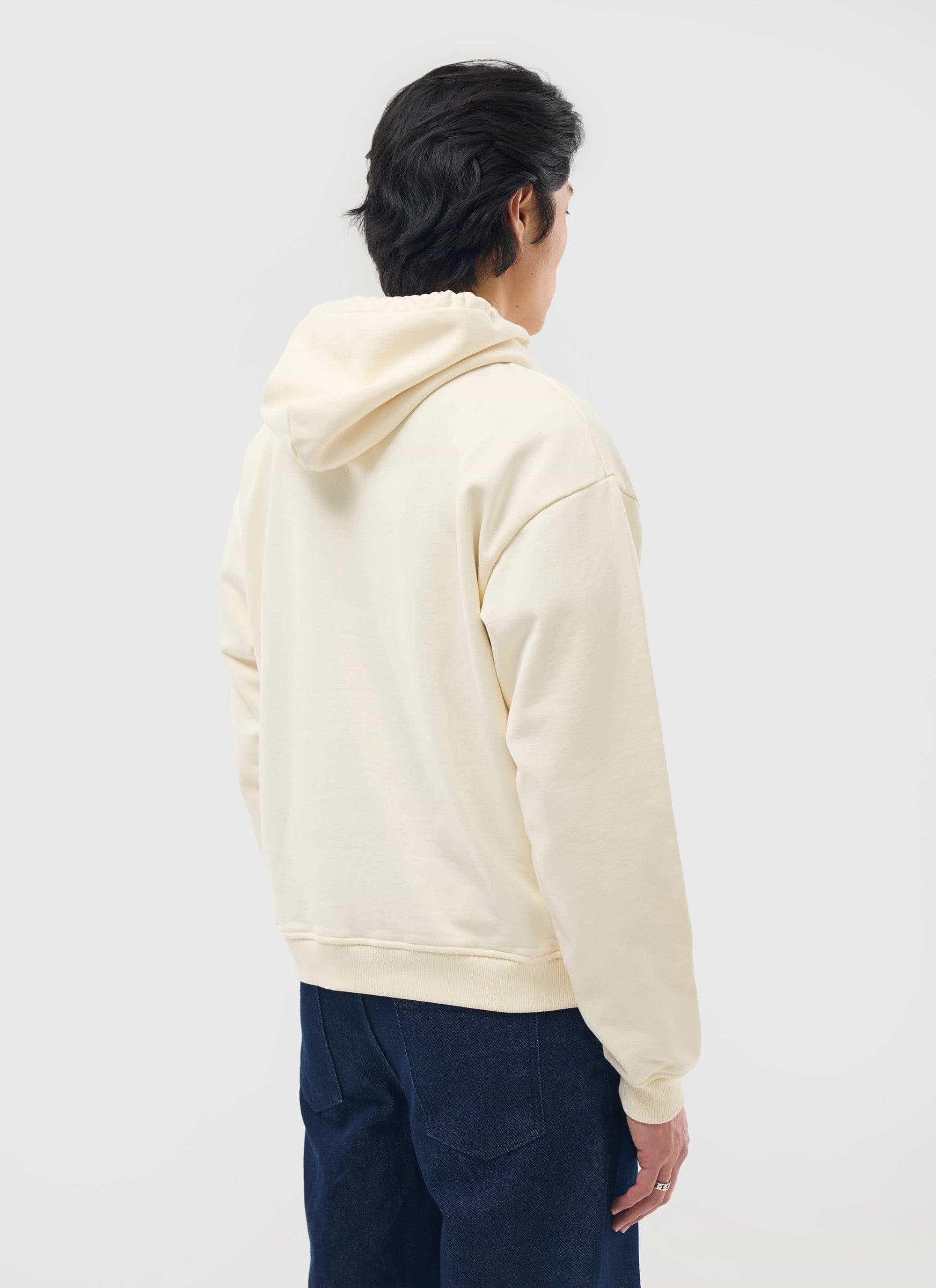 A model, seen from behind, wears the ecru Perci Chainstitch Hoodie from Percival with dark blue jeans.
