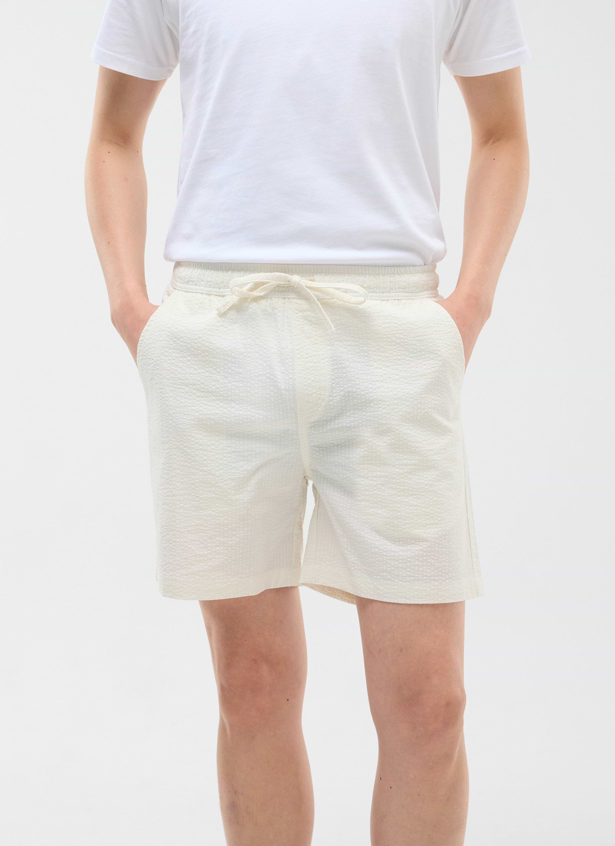 The Percival Classic Shorts in cream seersucker, shown on a model with a white t-shirt and hands in pockets.