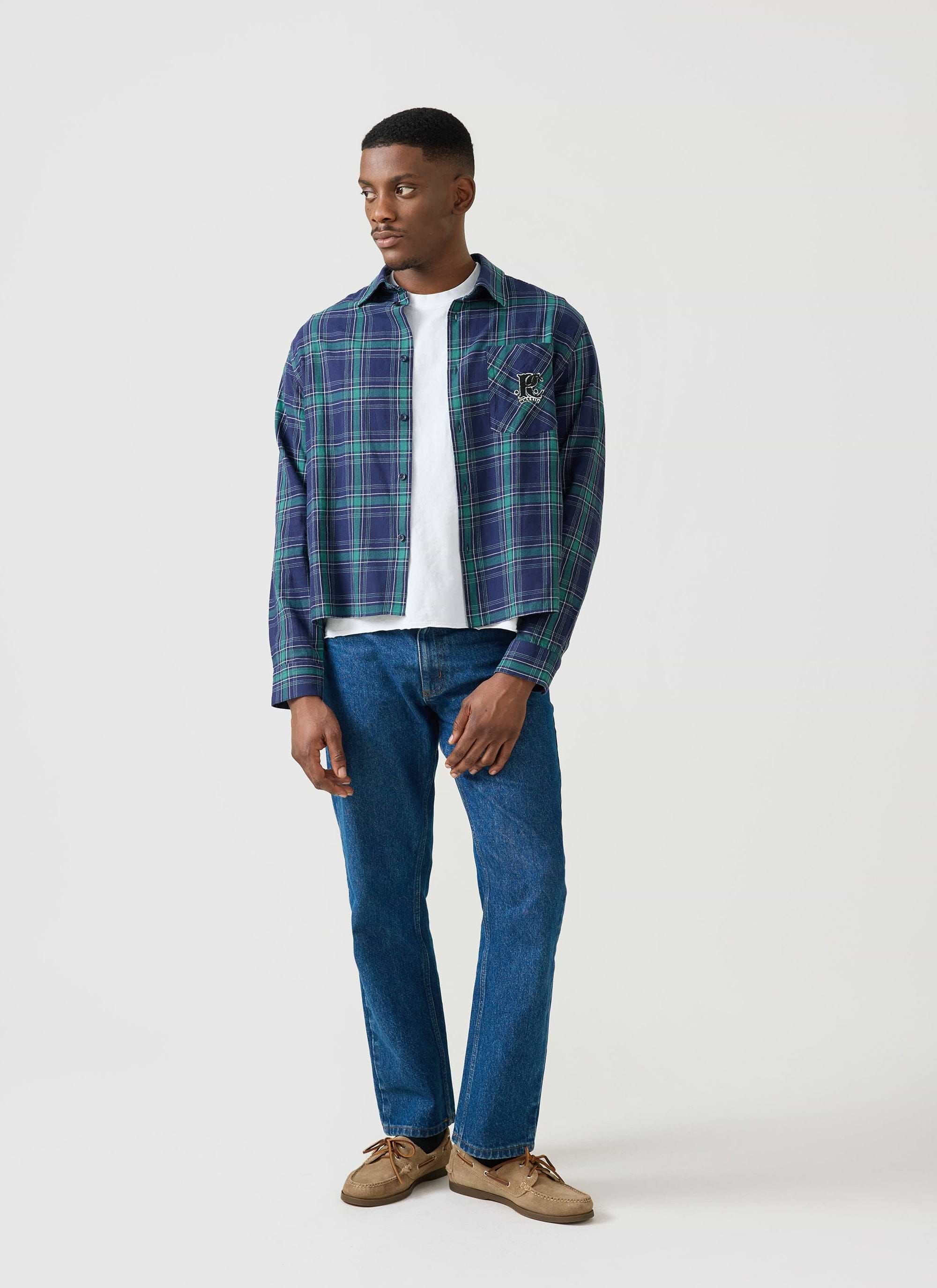A man models the Percival x Champion check boxy shirt, worn open over a white t-shirt with blue jeans.