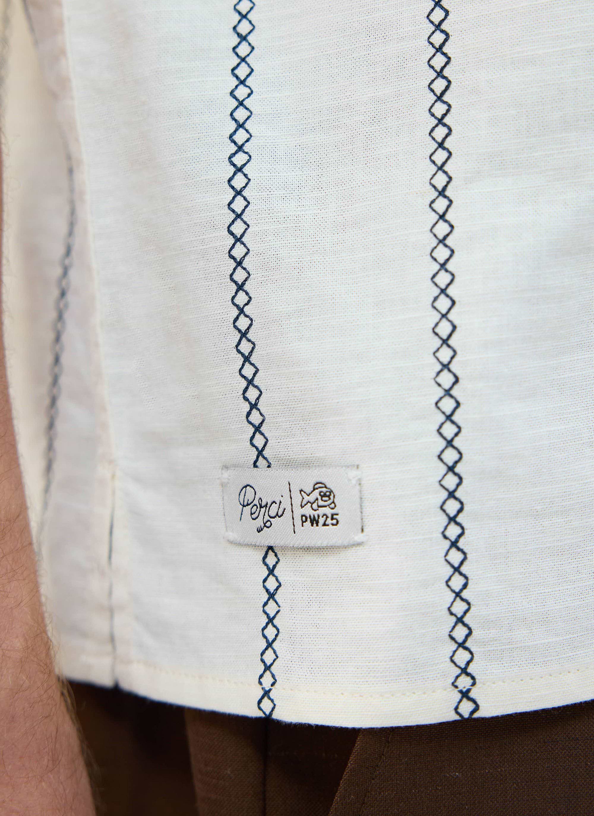 A close-up of the Percival Cross Stitch shirt in ecru, detailing the blue embroidery and signature 'Perci PW25' patch.