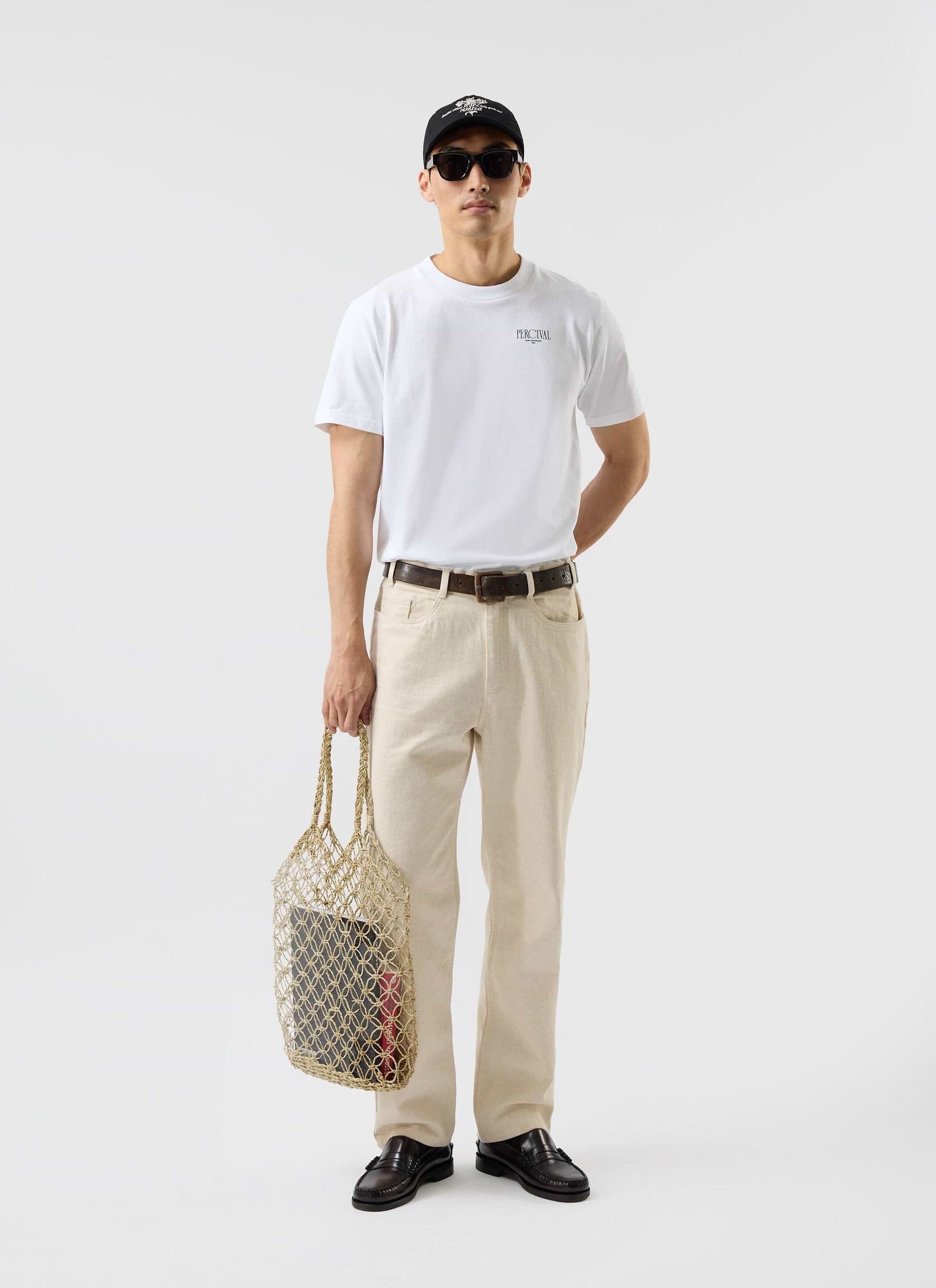 A model wears the white Percival Bistro T Shirt, styled with cream trousers, sunglasses, a cap, and holding a string bag.