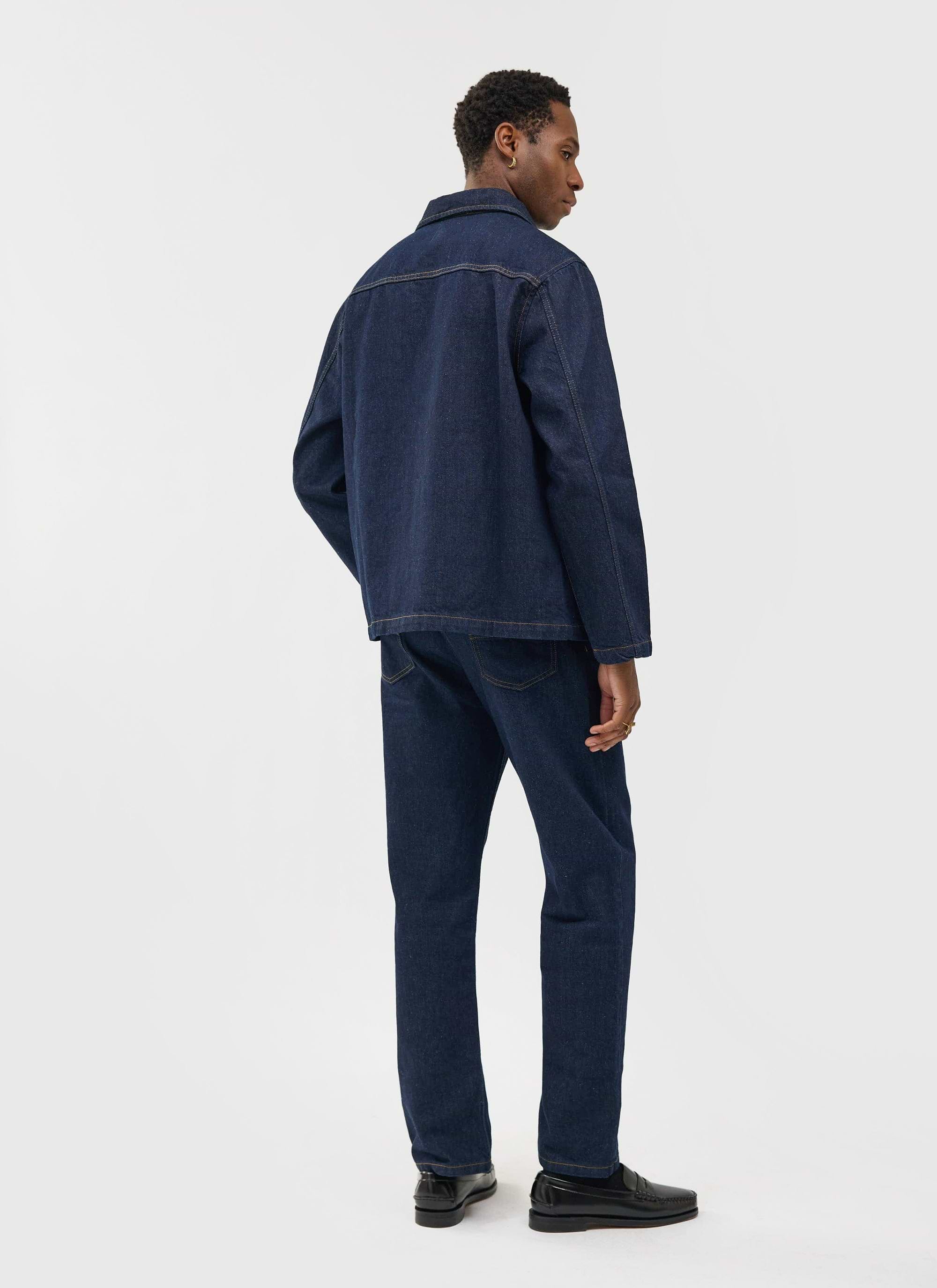 A model wears the Percival Gove Chore Jacket in dark indigo denim, styled with matching jeans and viewed from the back.