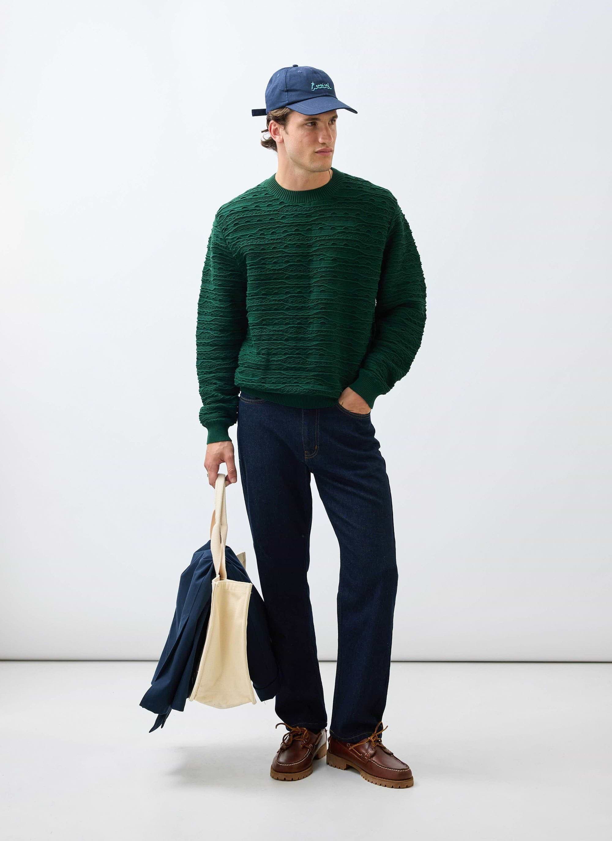 A model wears the Percival Tales Knitted Crew Neck Jumper in forest green styled with dark jeans, a blue cap, and a tote bag.