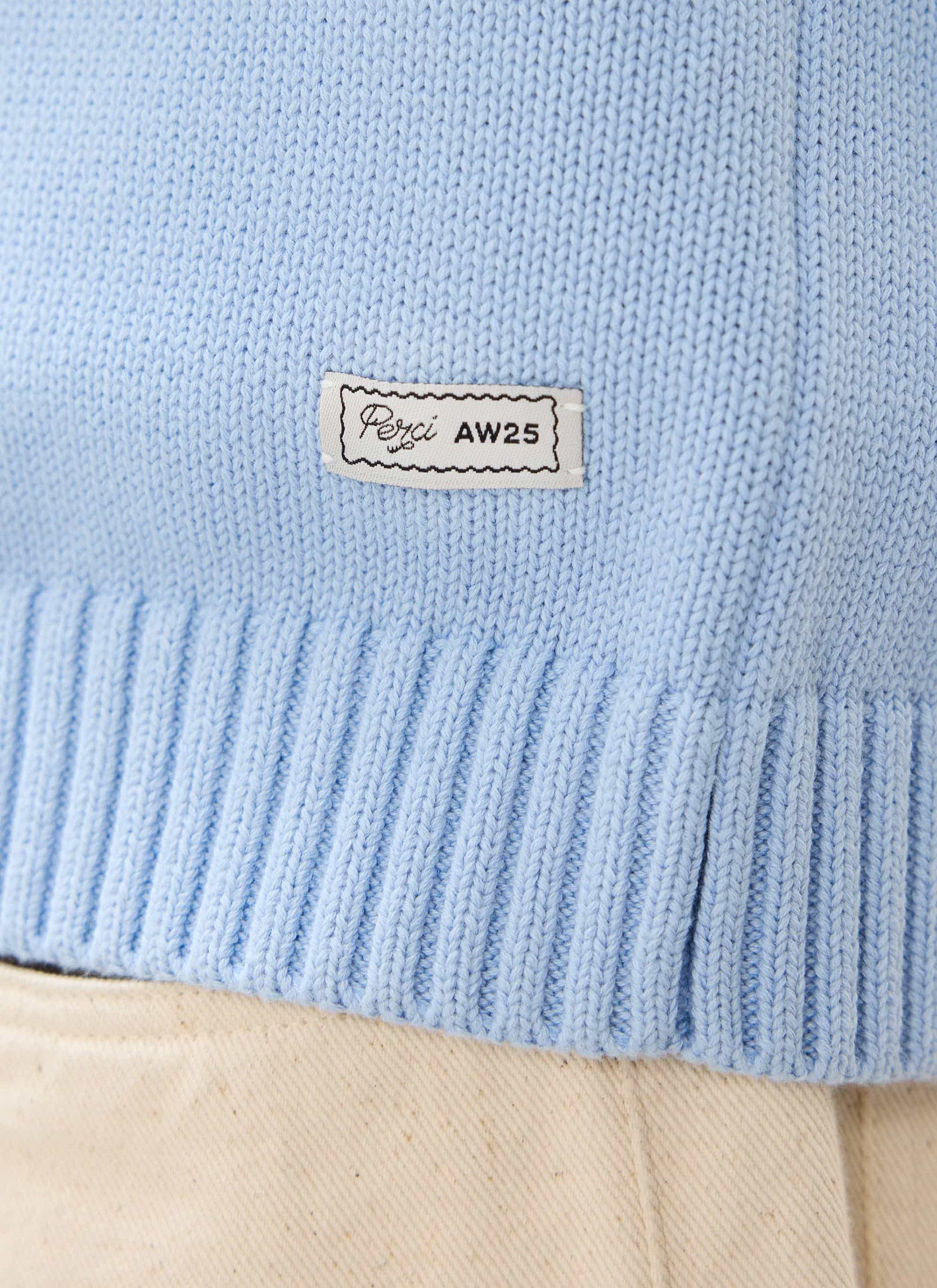 A close-up of the Percival Verso Short Sleeve Knitted Shirt in heather blue, showing the AW25 patch on the ribbed hem.