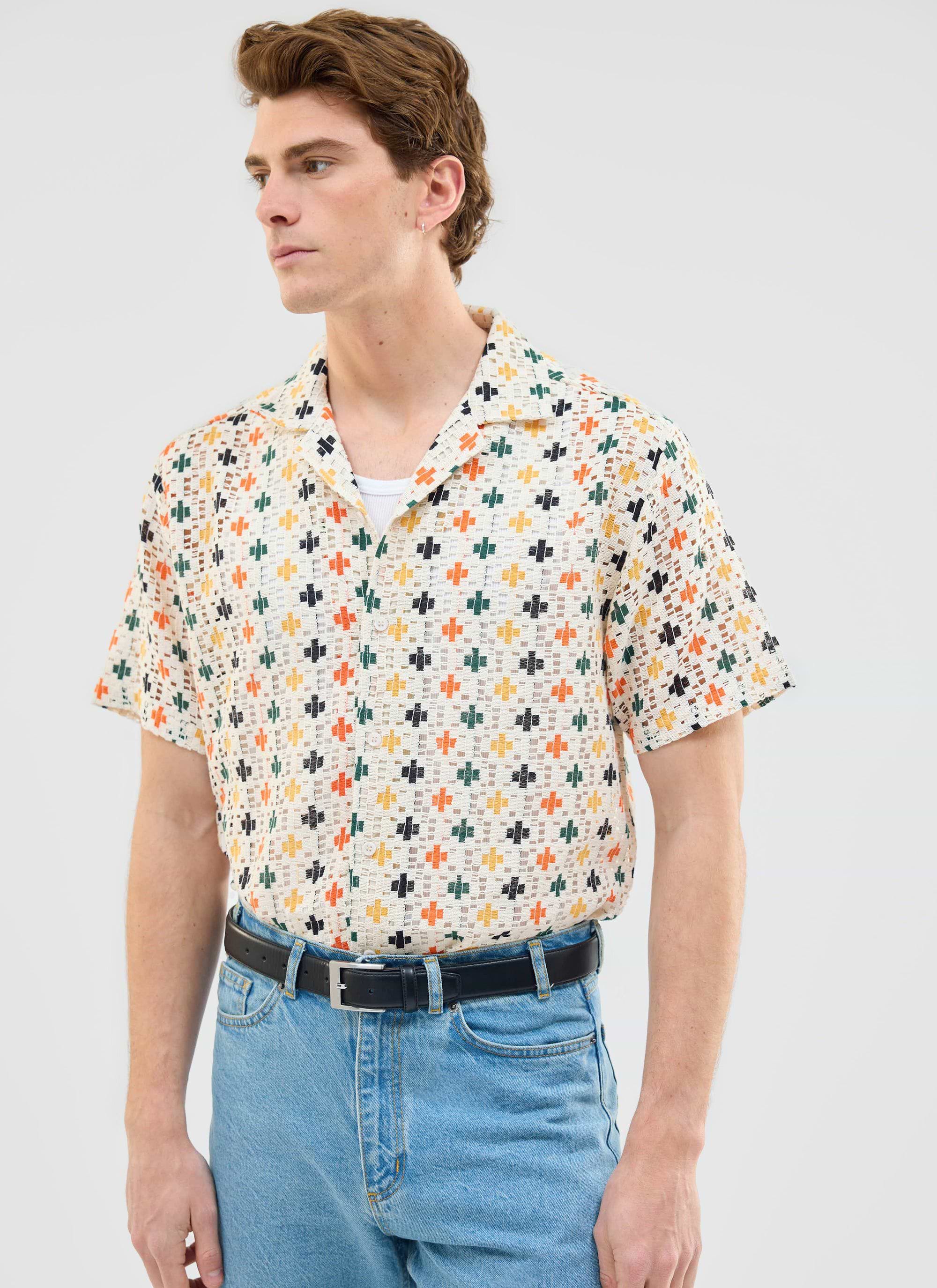 A model wears the Percival Bailing Crochet Cuban Shirt, an ecru shirt with a multi-coloured cross pattern, with blue jeans.