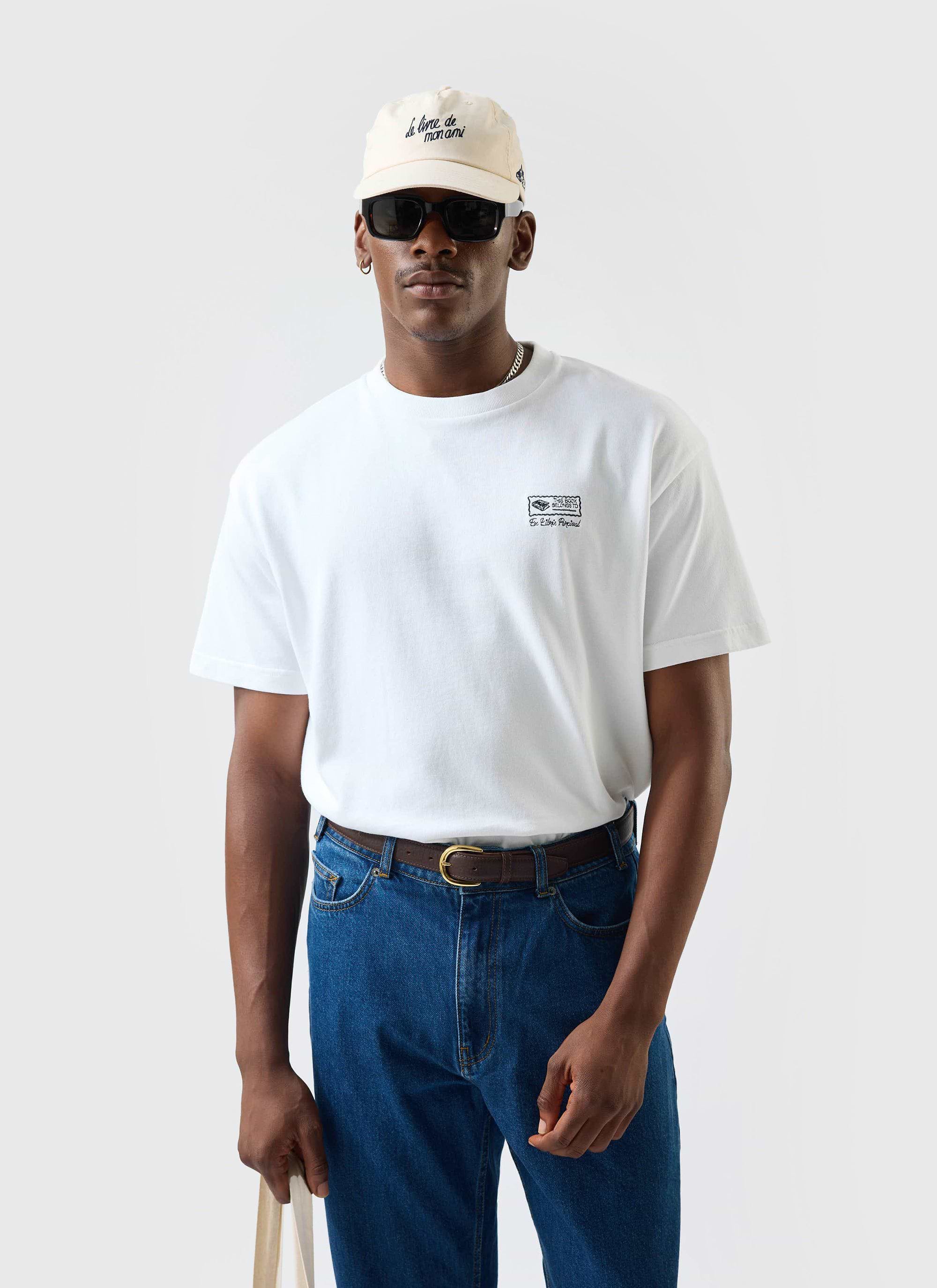 A man models the white Ex Libris oversized t-shirt from Percival, styled with blue jeans, sunglasses and a cream cap.