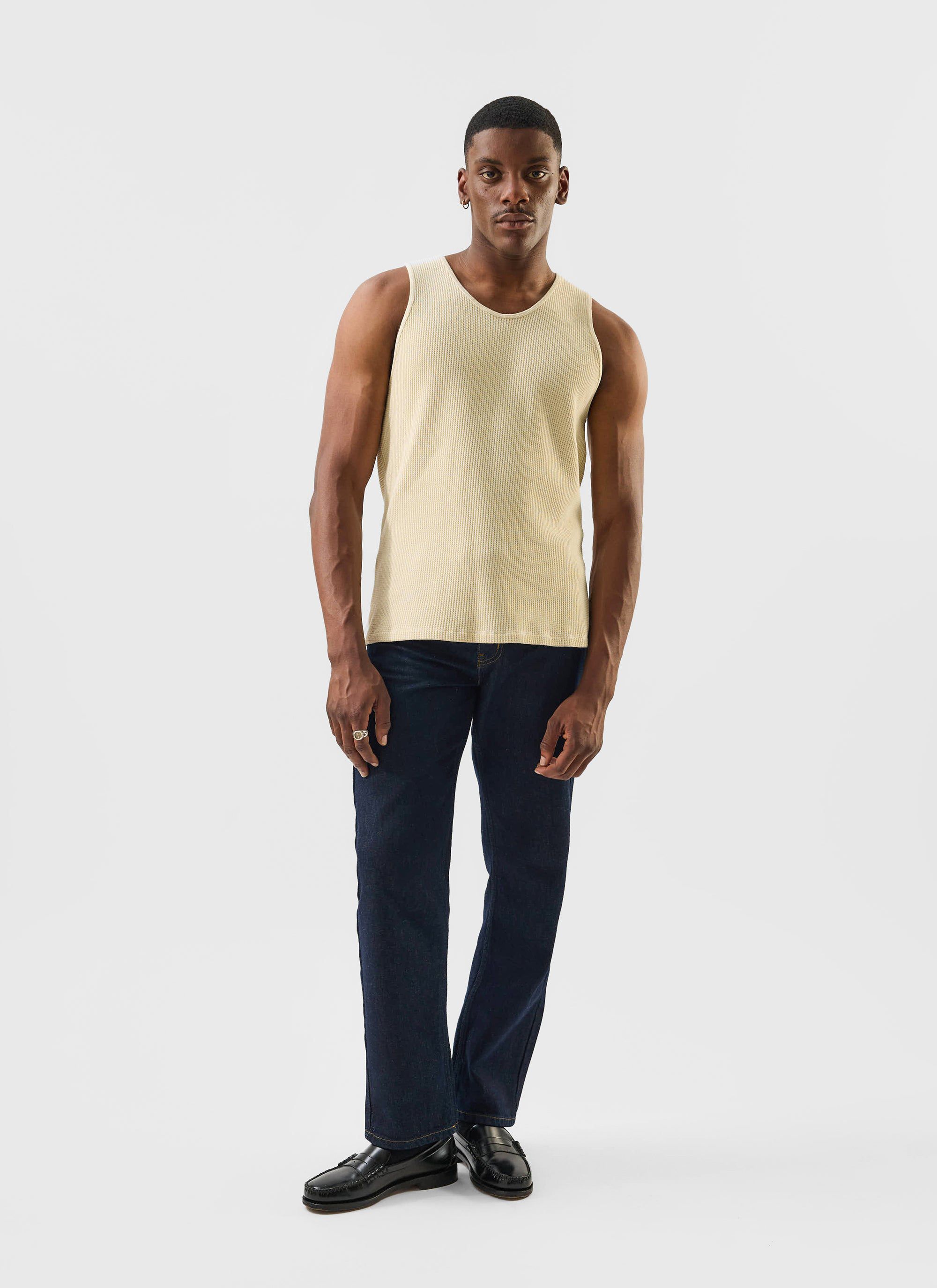 A man models the Percival Cotton Waffle Vest in ecru, styled with dark denim jeans and black loafers in a studio.