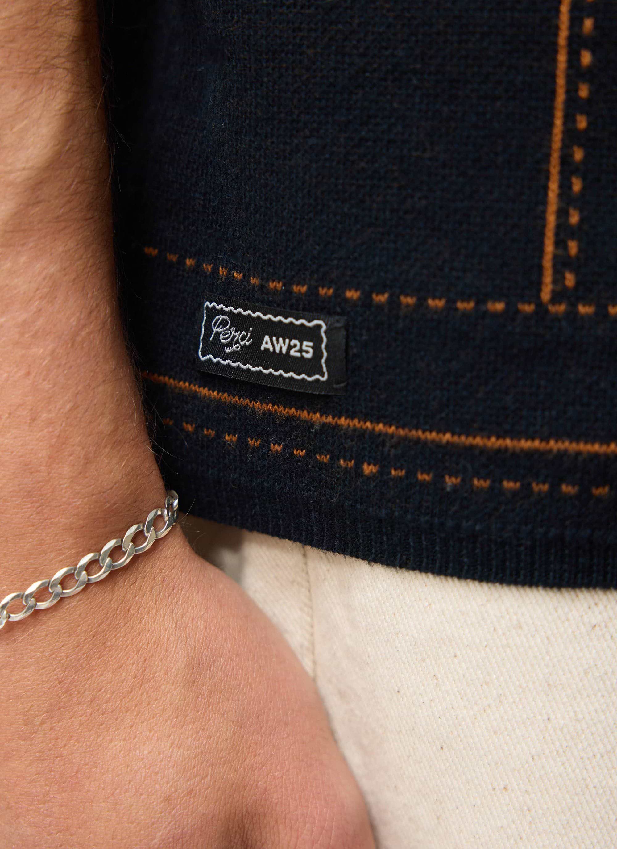 A close-up shot of the Percival Folio Knitted Shirt in dark navy, showing the jacquard detail and AW25 branded hem label.