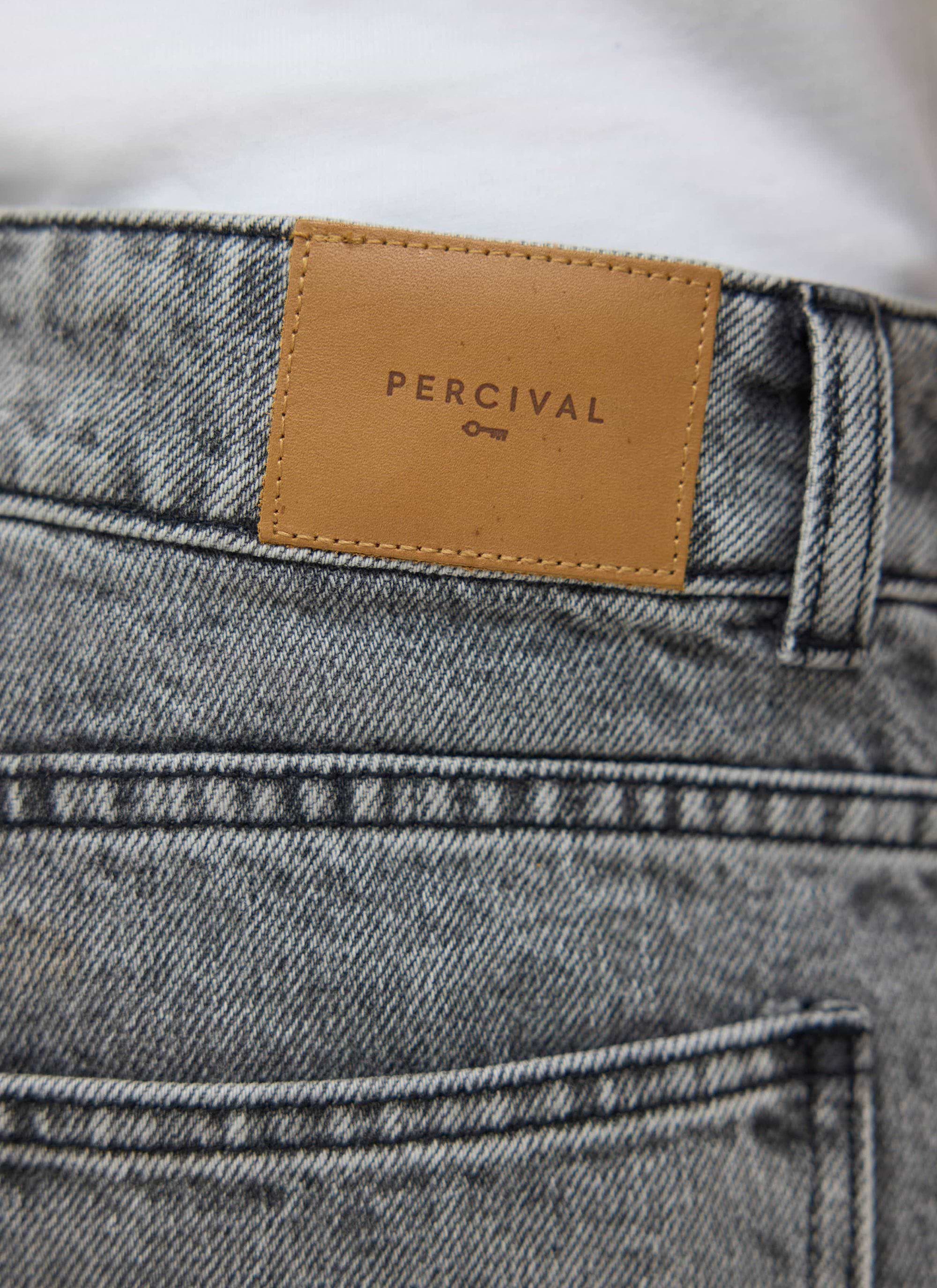A close-up of the Percival Wide Leg Denim Jeans in warm grey wash, focusing on the tan leather logo patch.