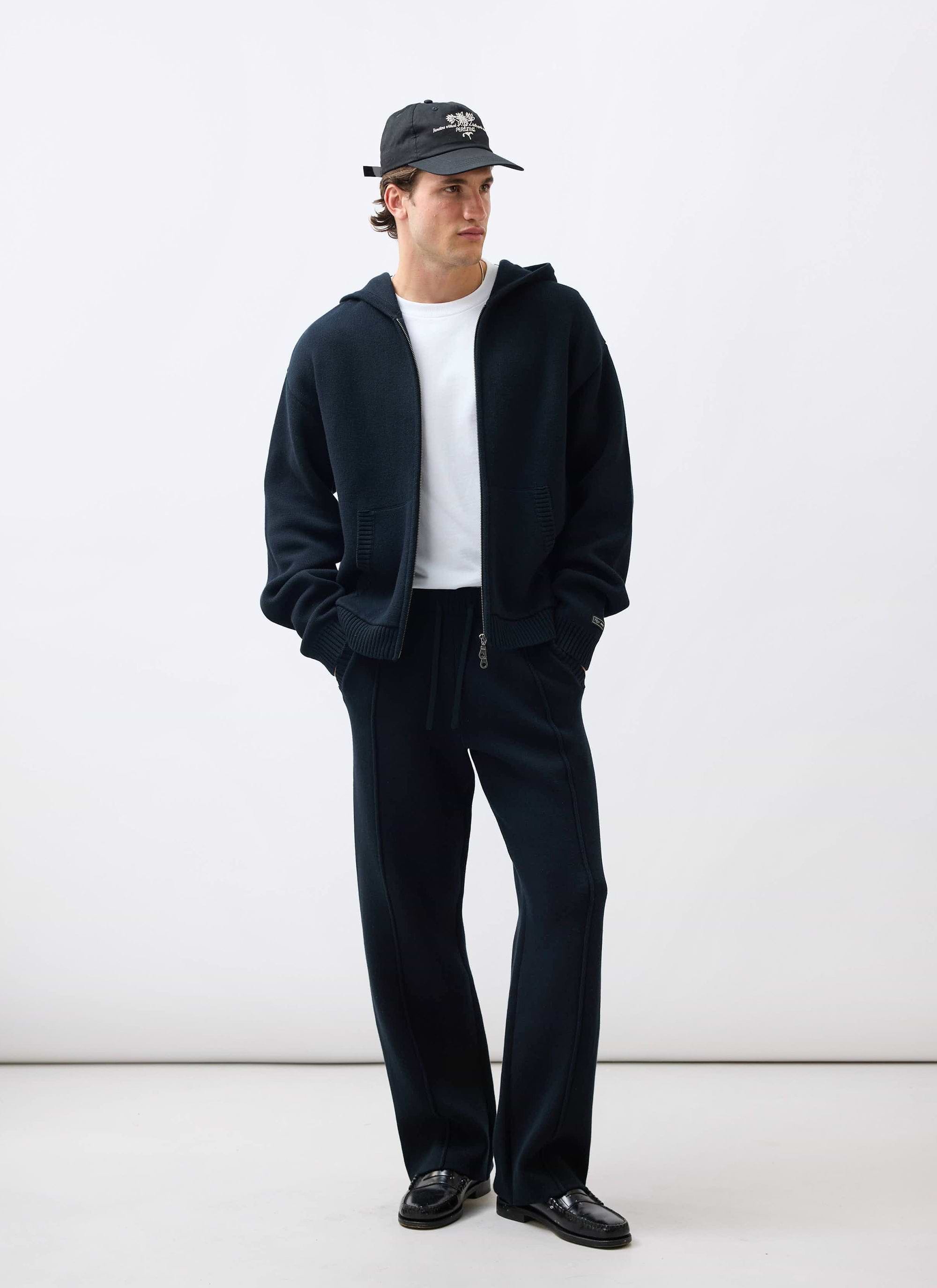 A model wears the black Percival Chronicle Knitted Hoodie and matching track pants, styled with a white tee and a cap.