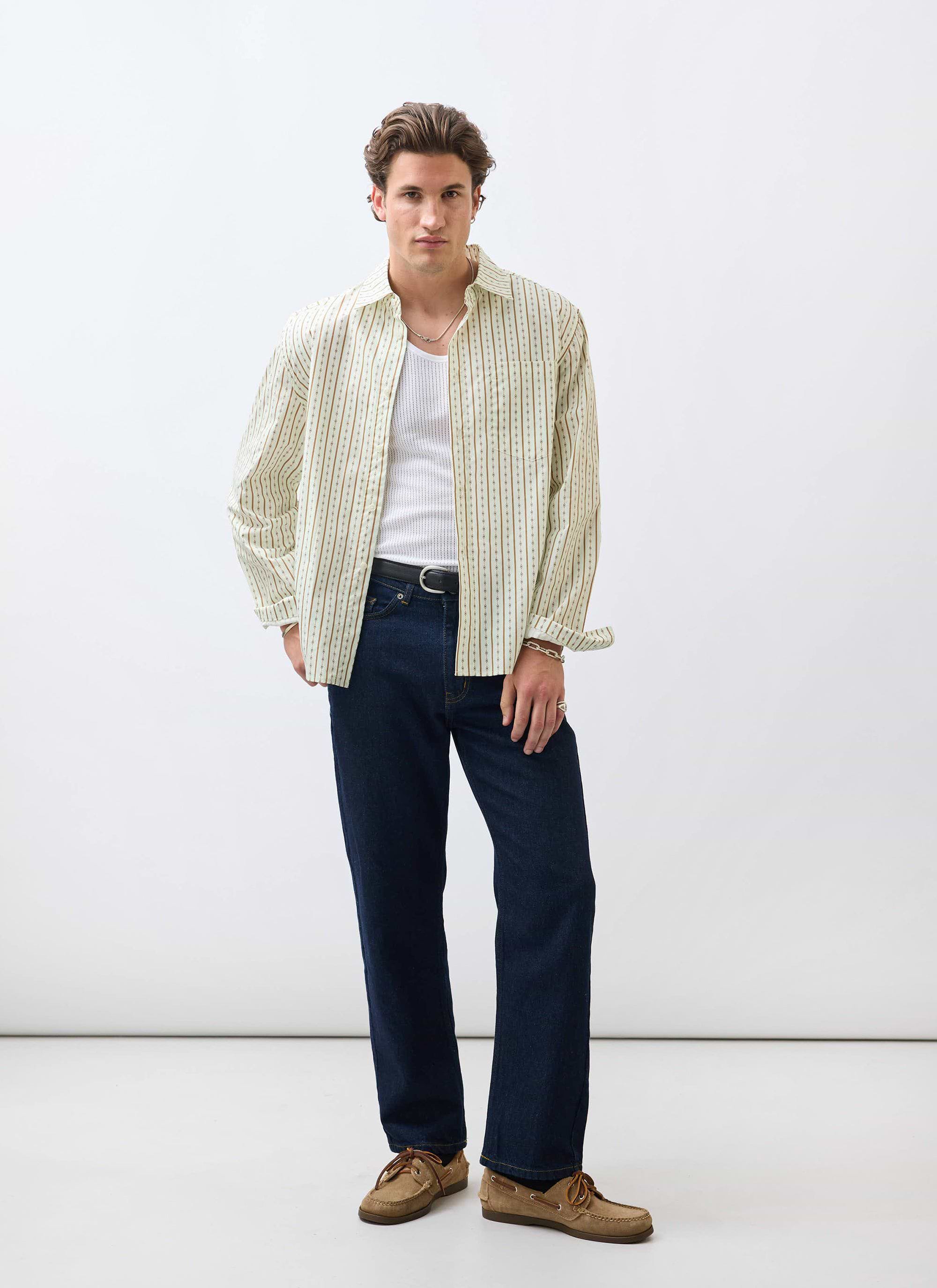 A model wears the Percival Binding Oversized Pearce Shirt in ecru, styled open over a white vest with dark denim jeans.