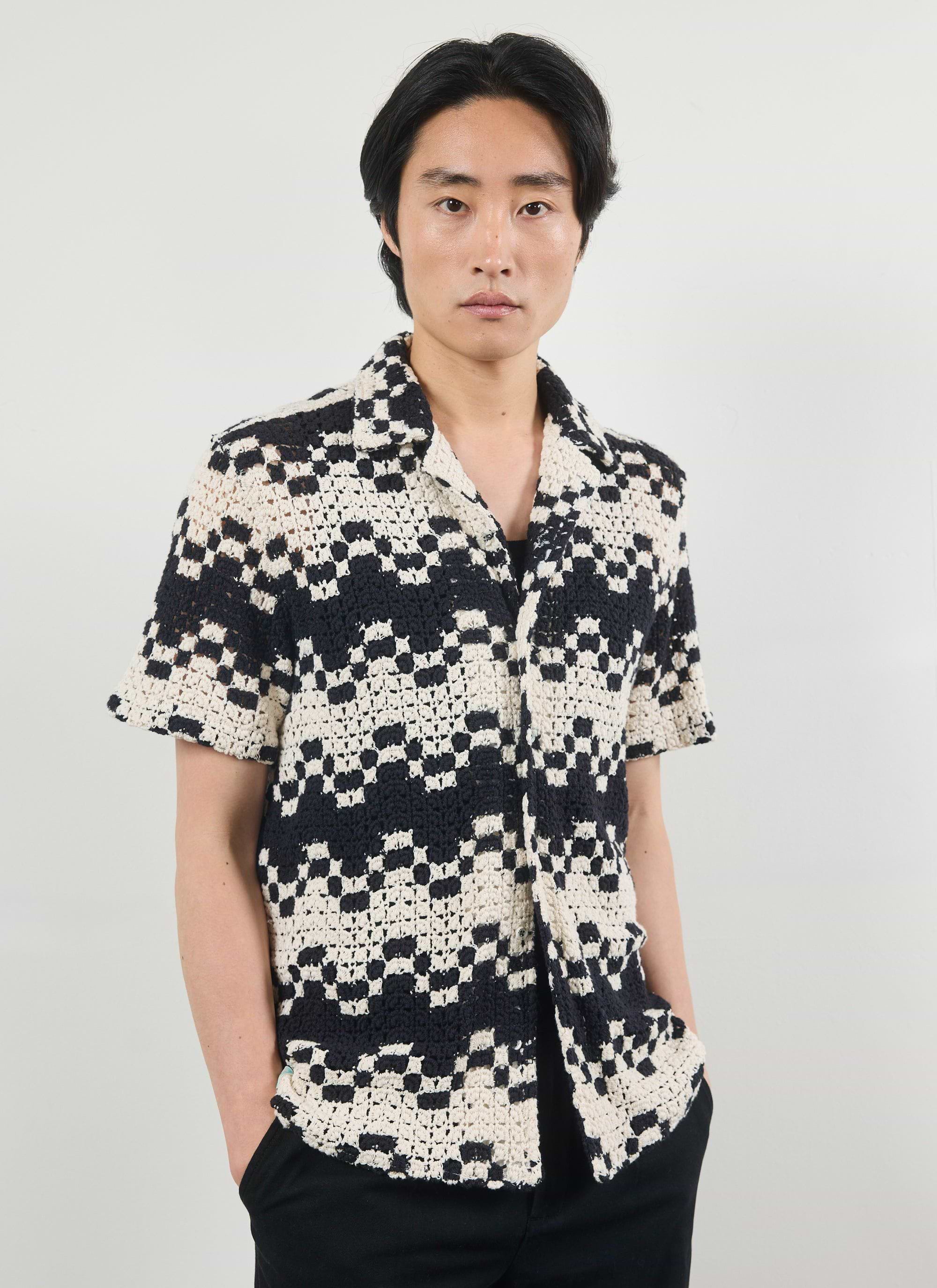 A man models the Percival Naples Crochet Cuban Shirt in an ecru and black geometric crochet knit against a white background.
