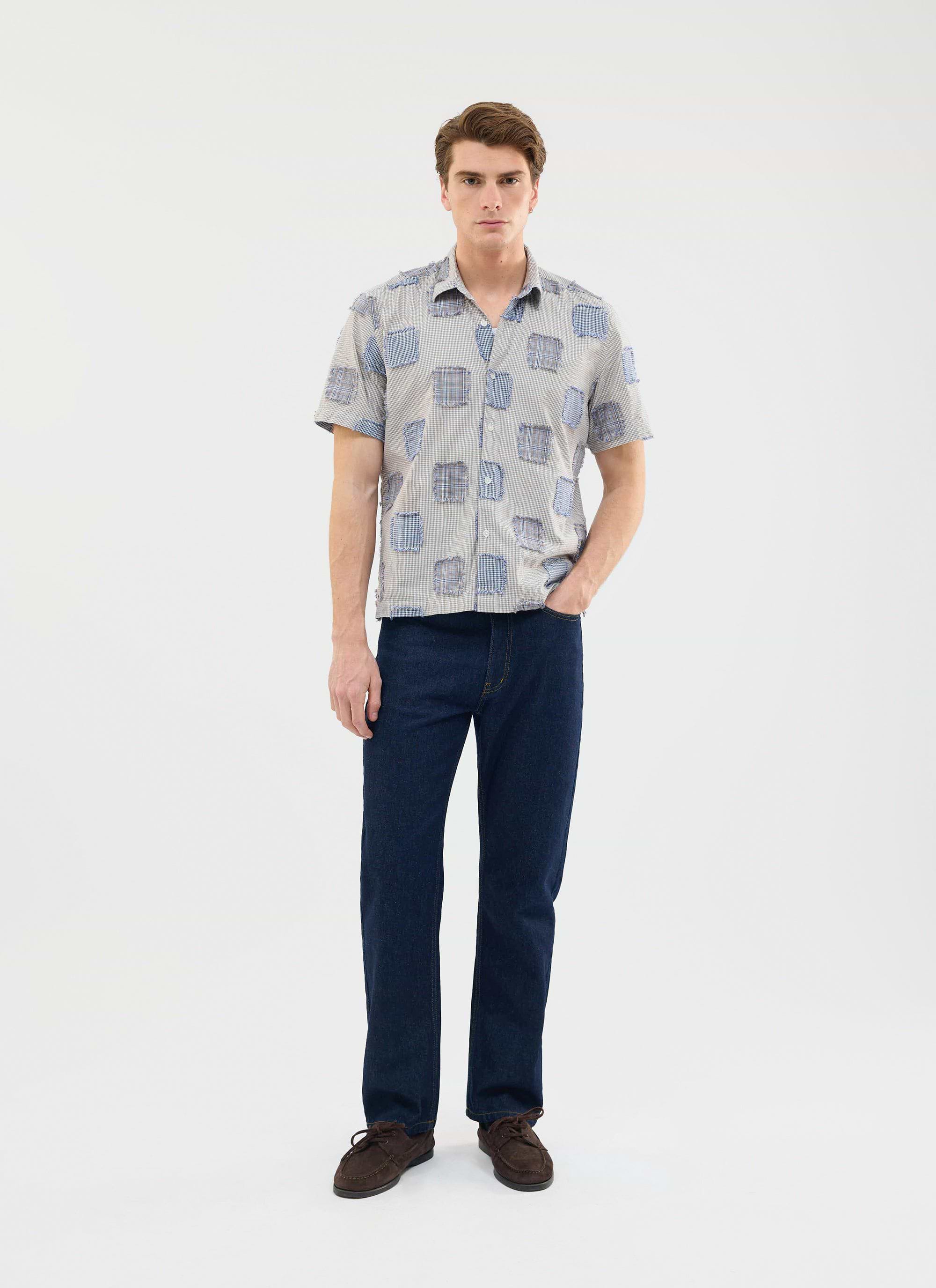 A man models the Percival Bruno Boxy Clerk Shirt in grey, featuring a patchwork design, styled with dark wash jeans.