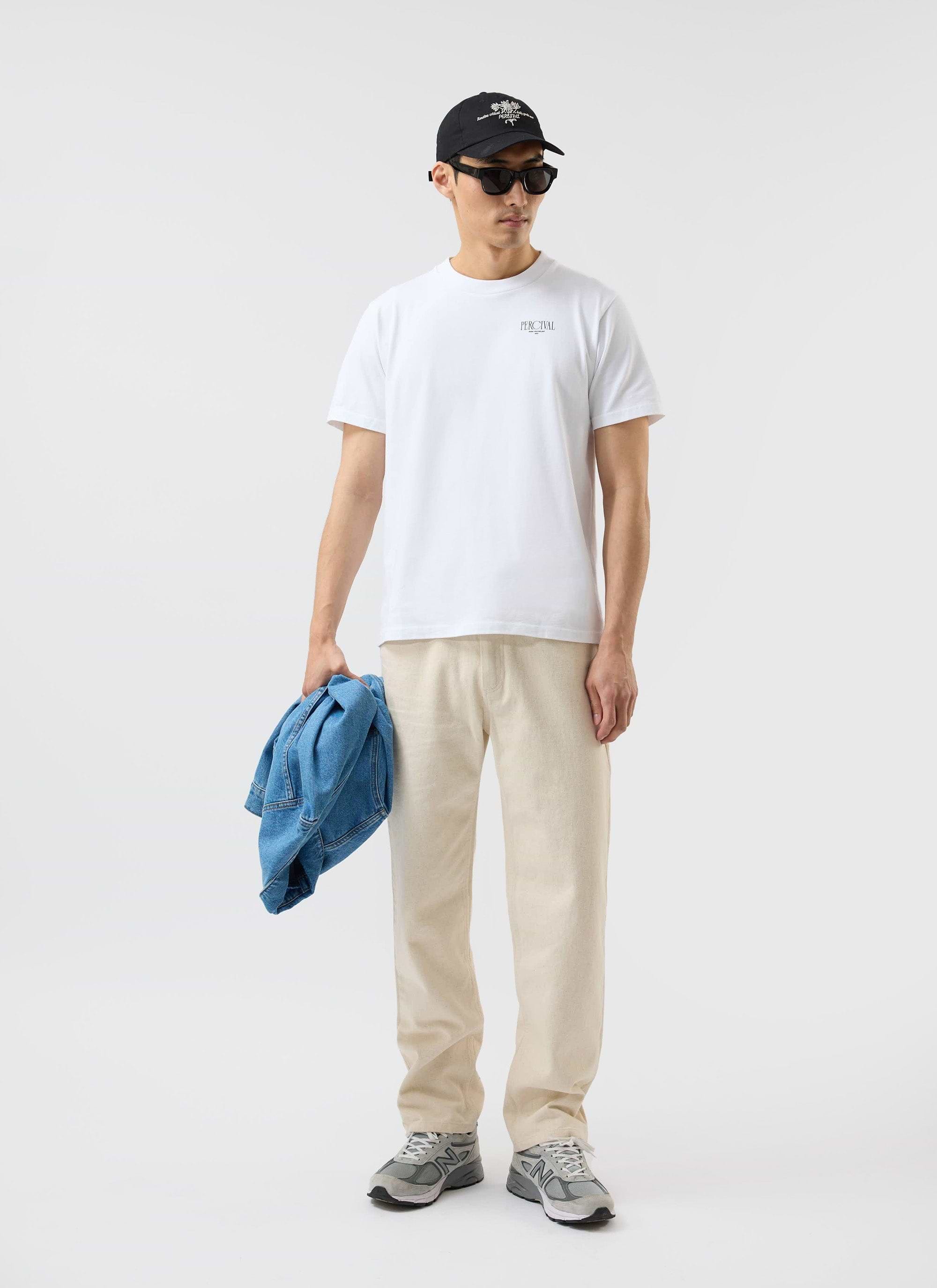 A model wears the white Percival Bistro T Shirt, styled with cream trousers, sunglasses and cap against a white background.