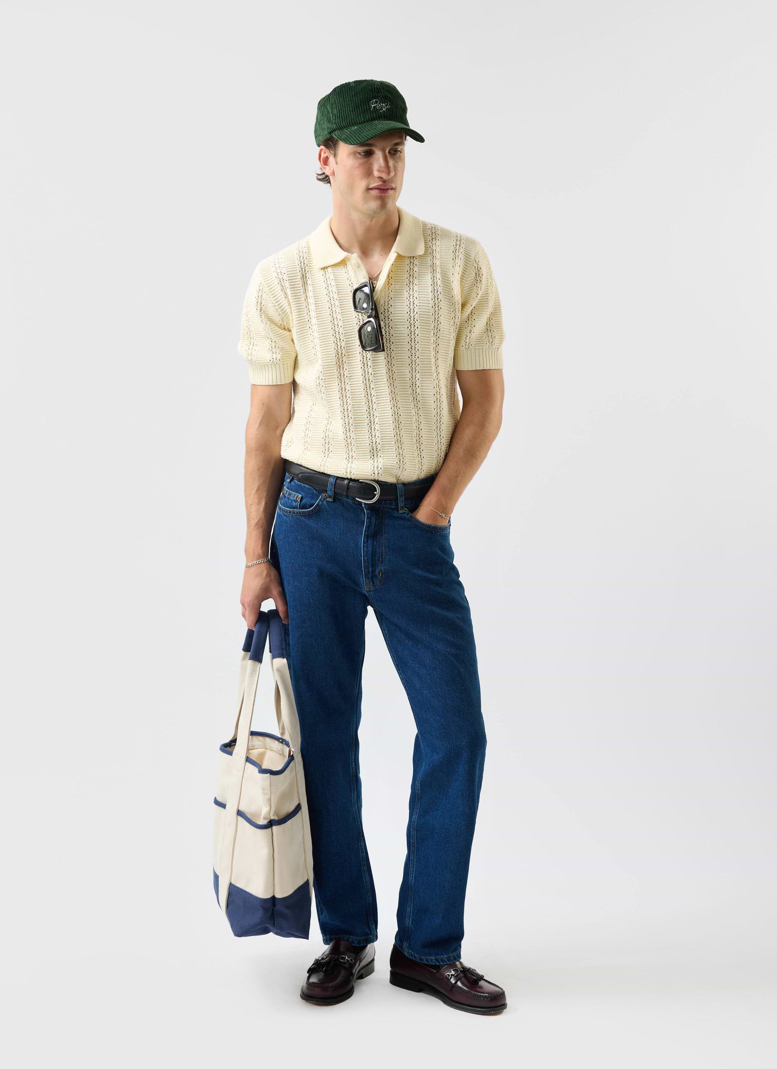A model styled in the Percival Addendum Knitted Polo Shirt in ecru cream, paired with blue jeans and a green corduroy cap.