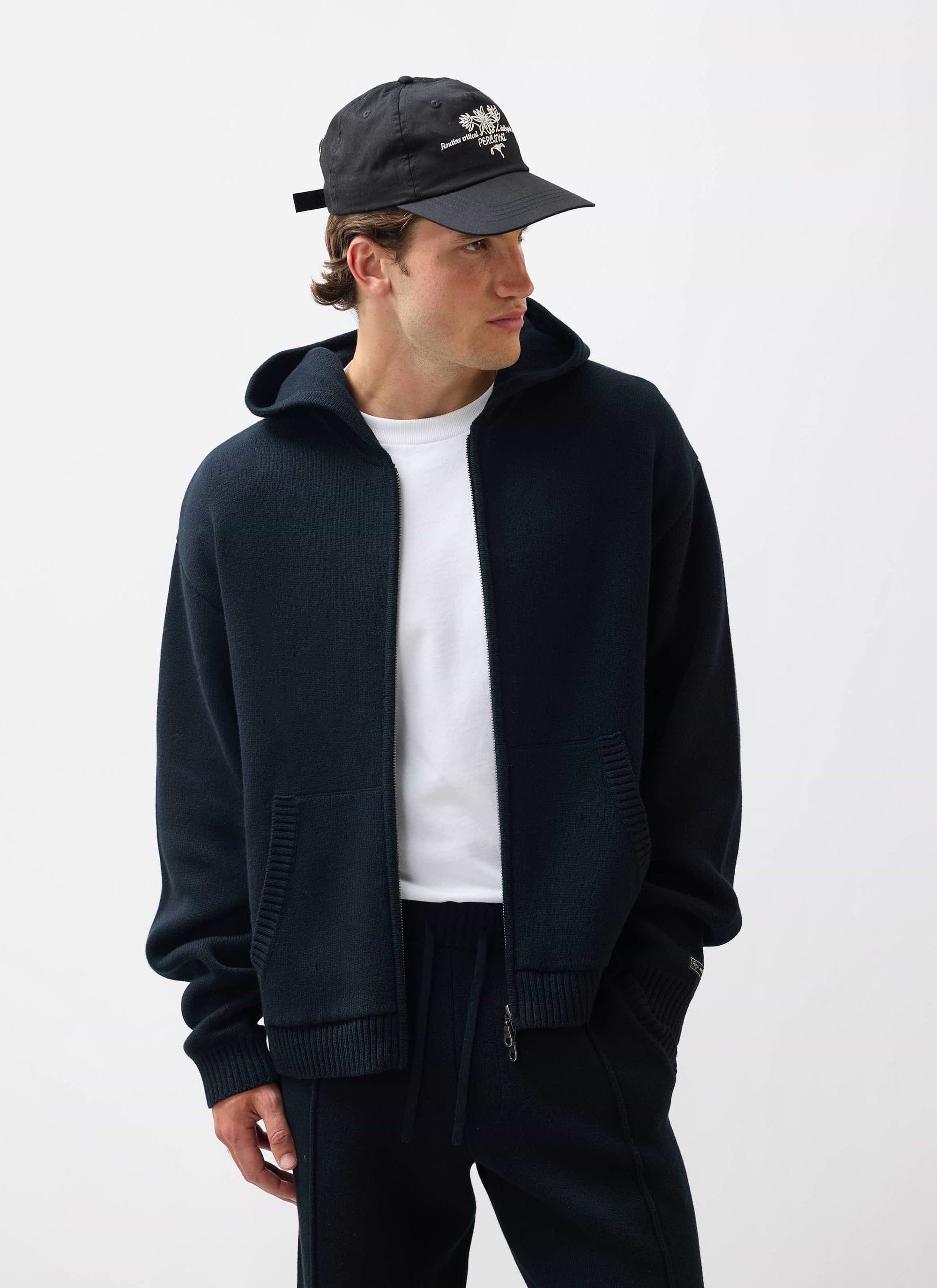 A model wears the black Percival Chronicle Knitted Hoodie, open over a white t-shirt, against a white studio background.
