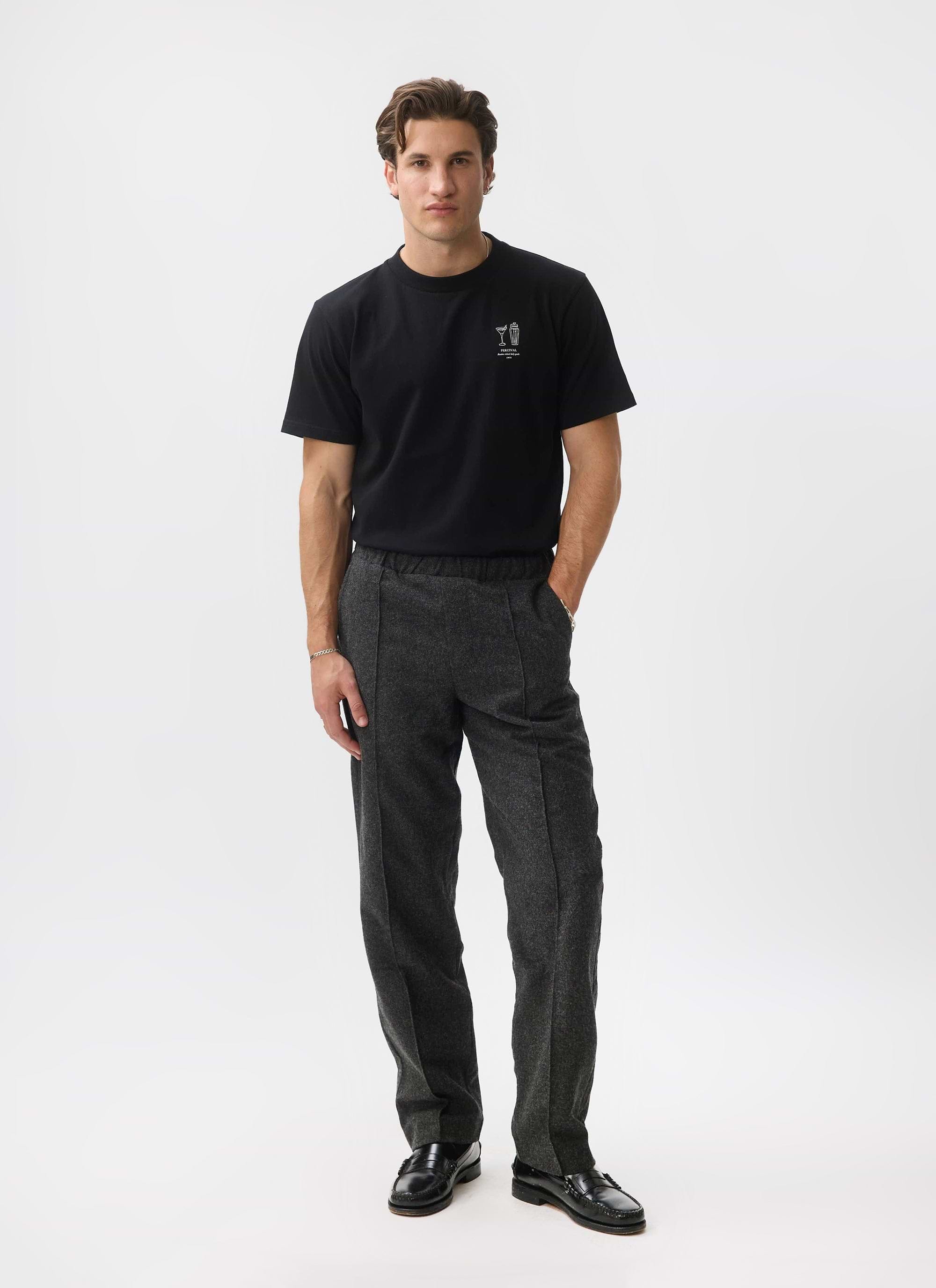 A model wears the Percival Caxton Smart Trousers in black brushed wool, styled with a black t-shirt and loafers.
