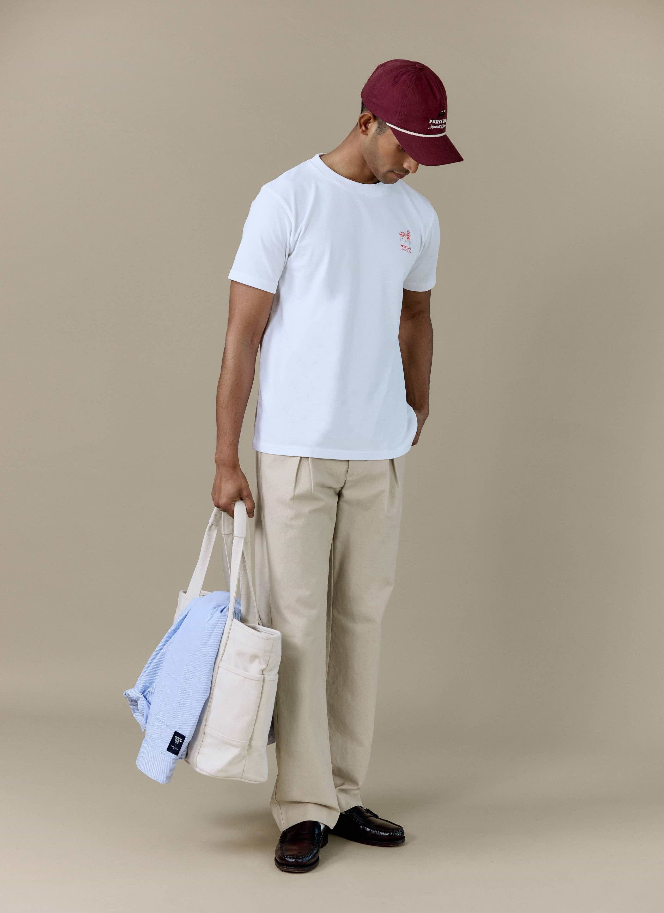 A model wearing the white Perci Fine Wines T Shirt by Percival, styled with beige trousers and a burgundy cap.