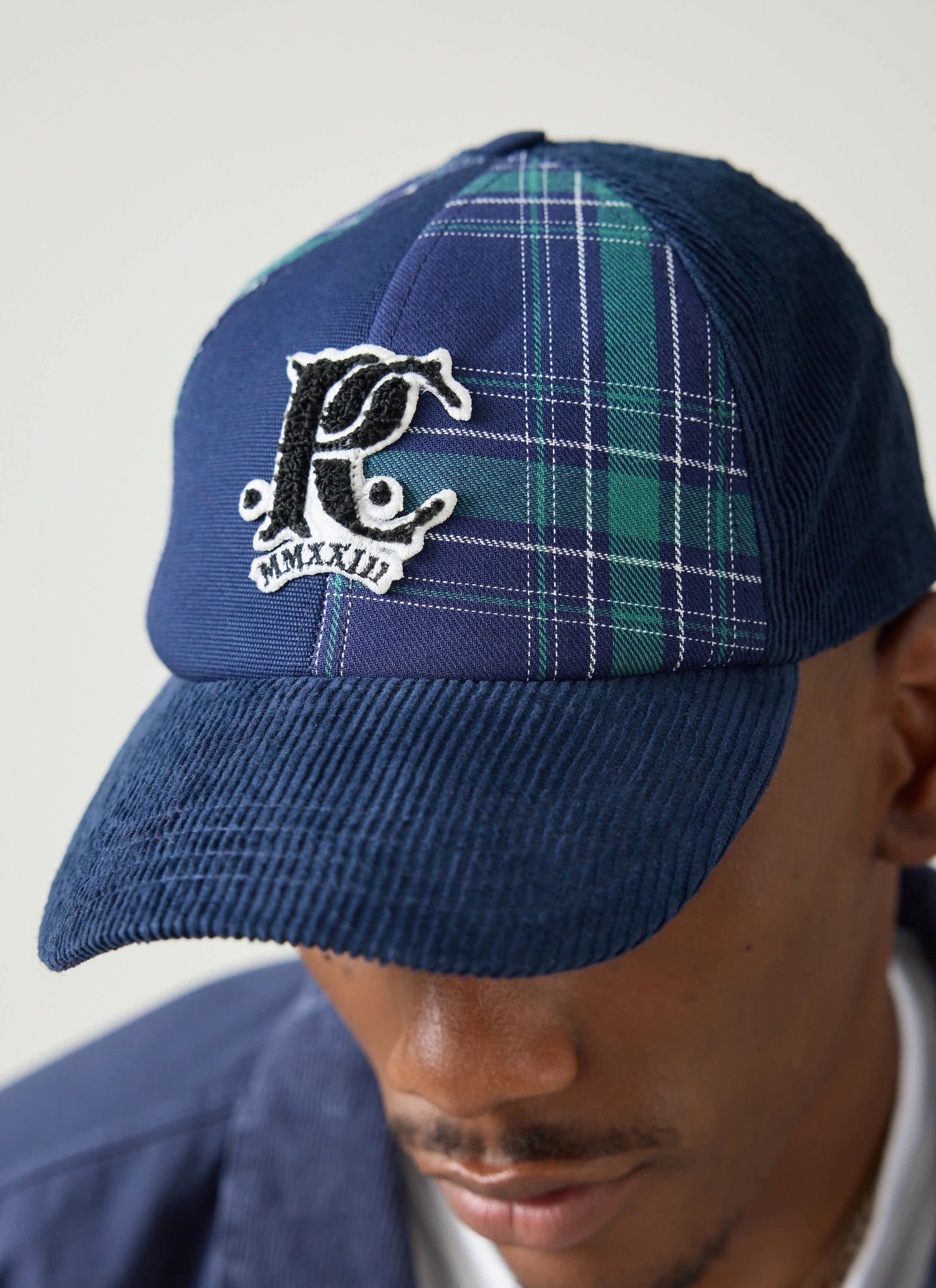 A close-up of a model wearing the Champion x Percival Cut and Sew Cap, detailing its navy corduroy and tartan panels.