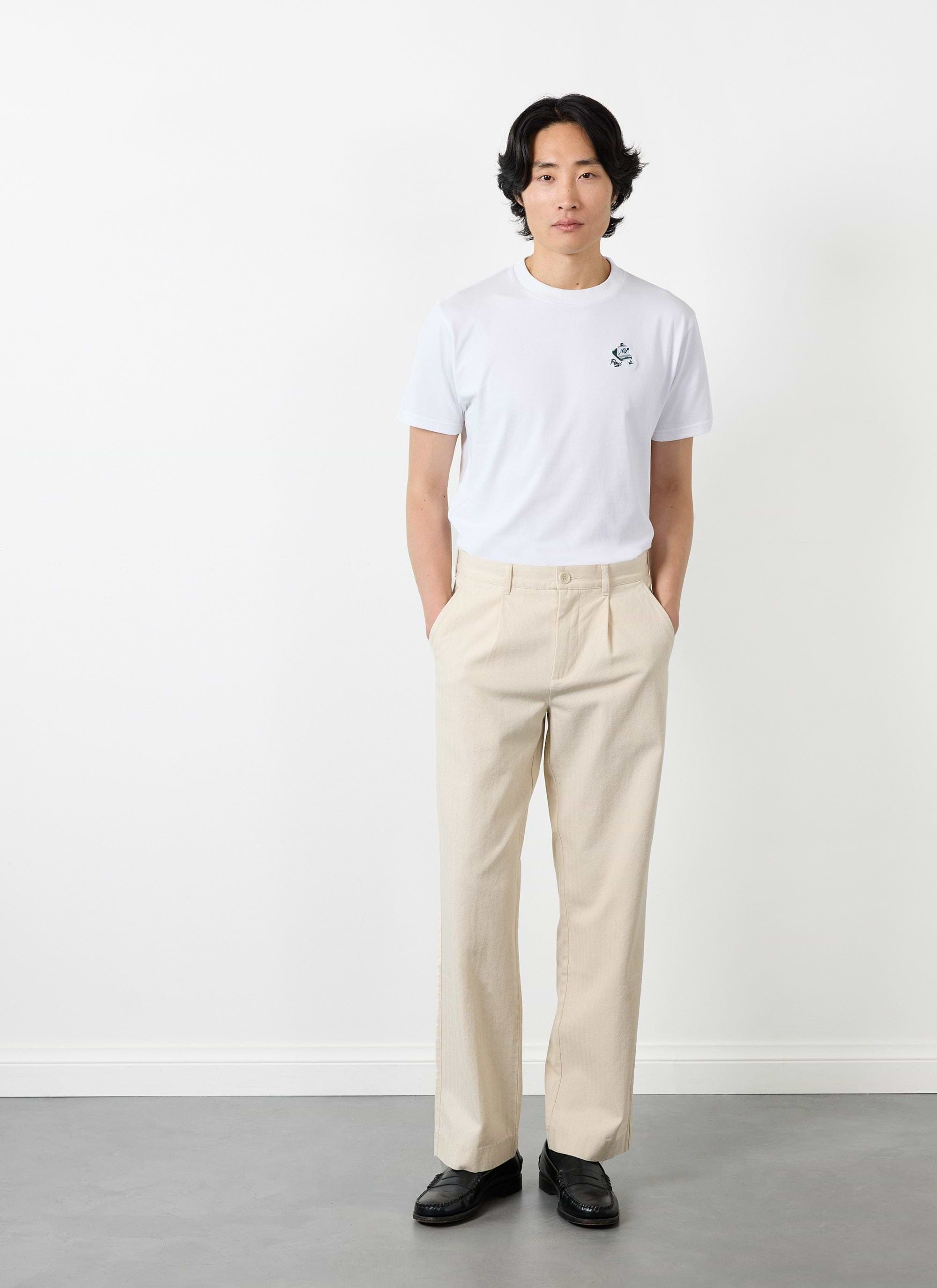 A model wears Percival's Program Pleated Trousers in ecru with a white t-shirt and black loafers.