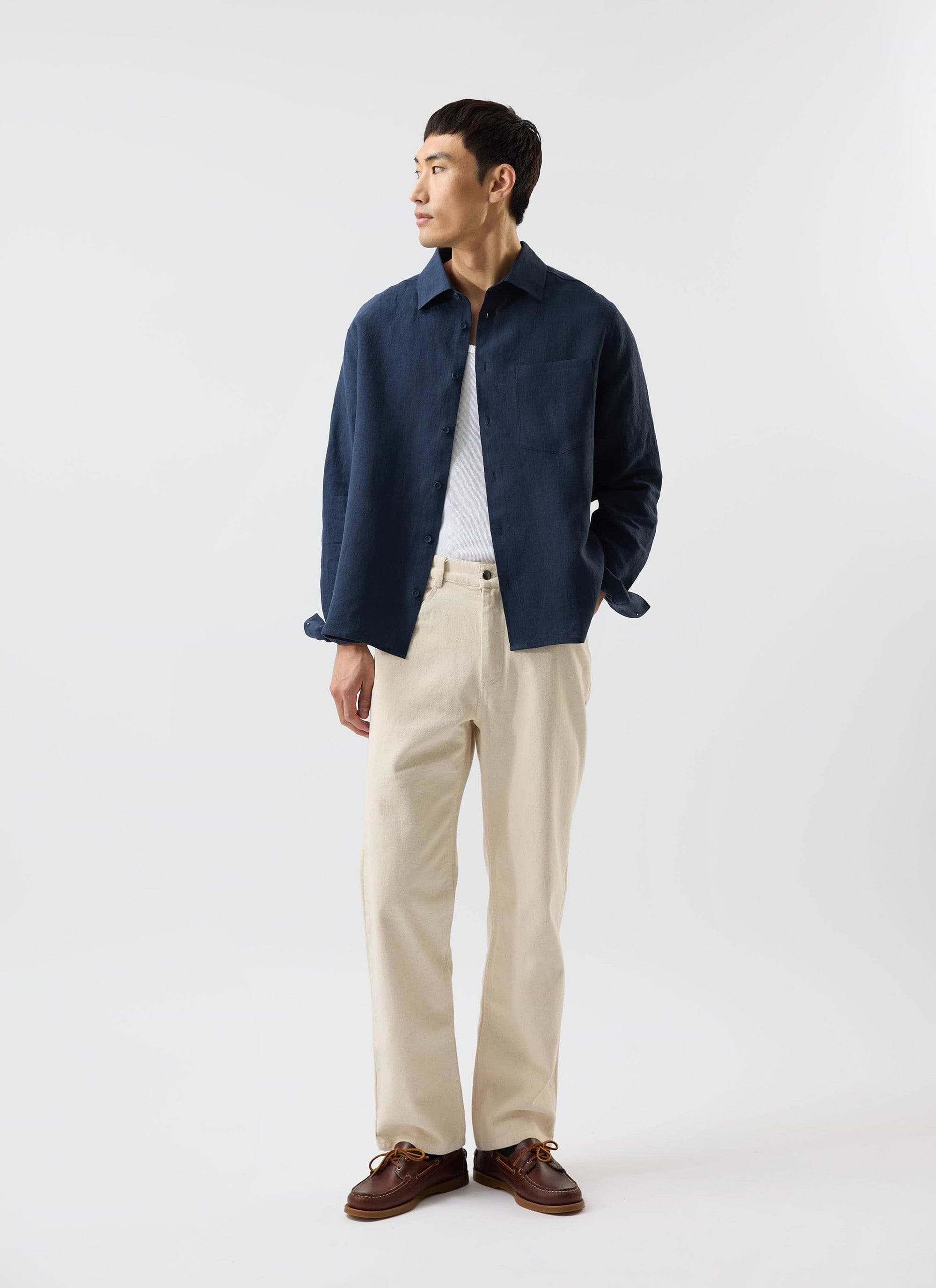 A man models the navy linen Pearce Shirt by Percival, open over a white t-shirt and paired with light-coloured trousers.