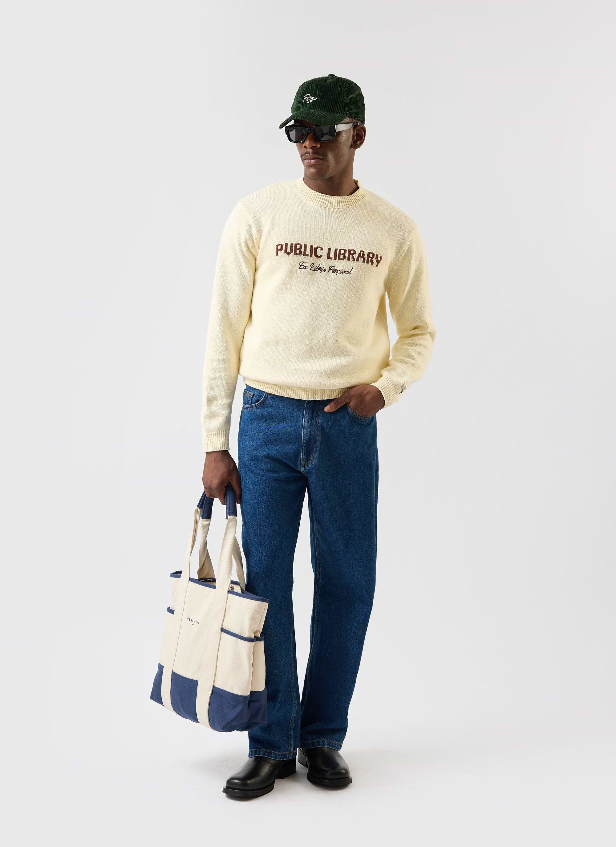 A model wears the ecru cream Public Library knitted jumper from Percival, styled with blue jeans, a tote bag and a green cap.