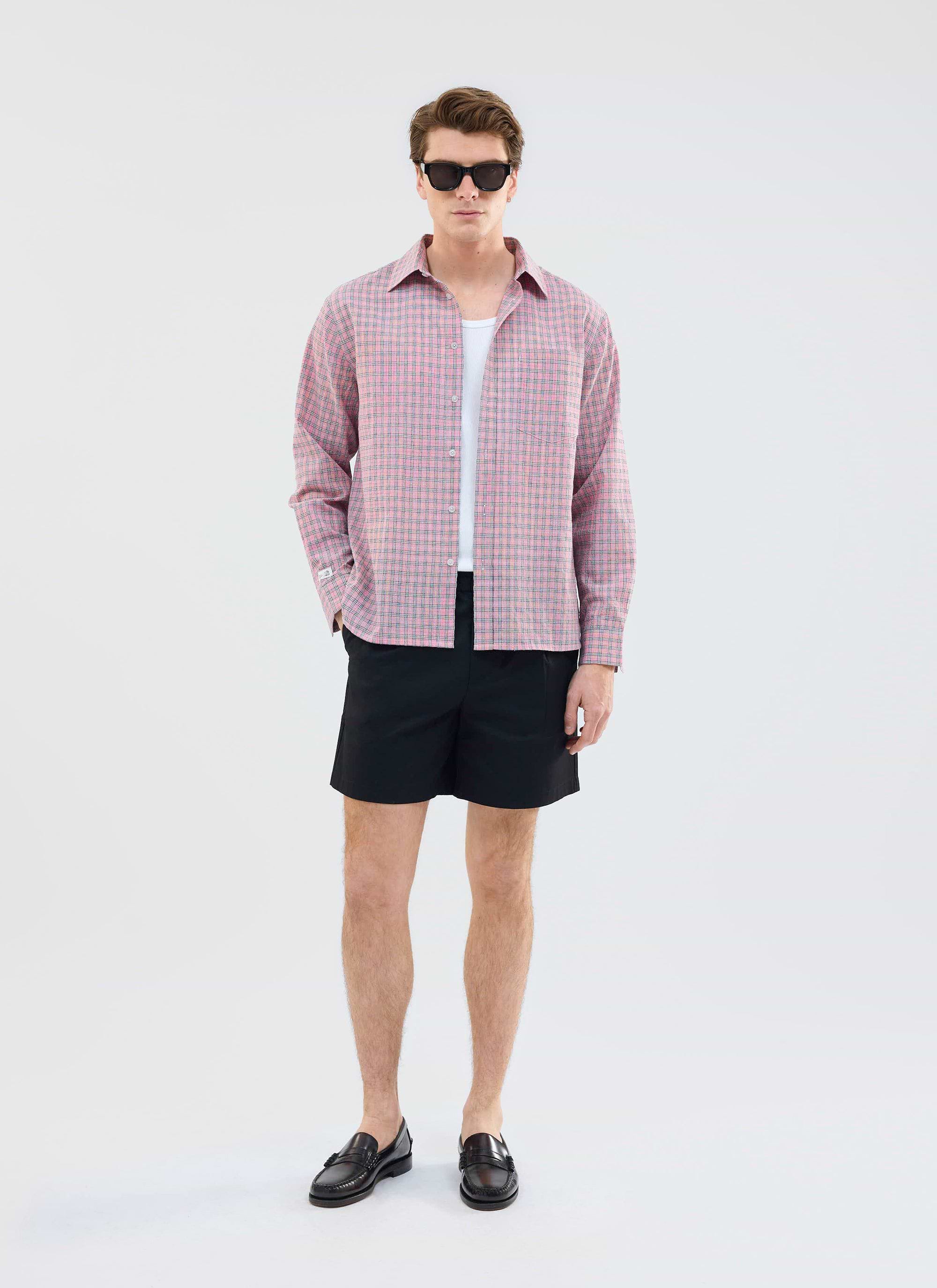 A model styles the black Percival Enzo Tailored Shorts with an open pink checked shirt, white vest, and black loafers.