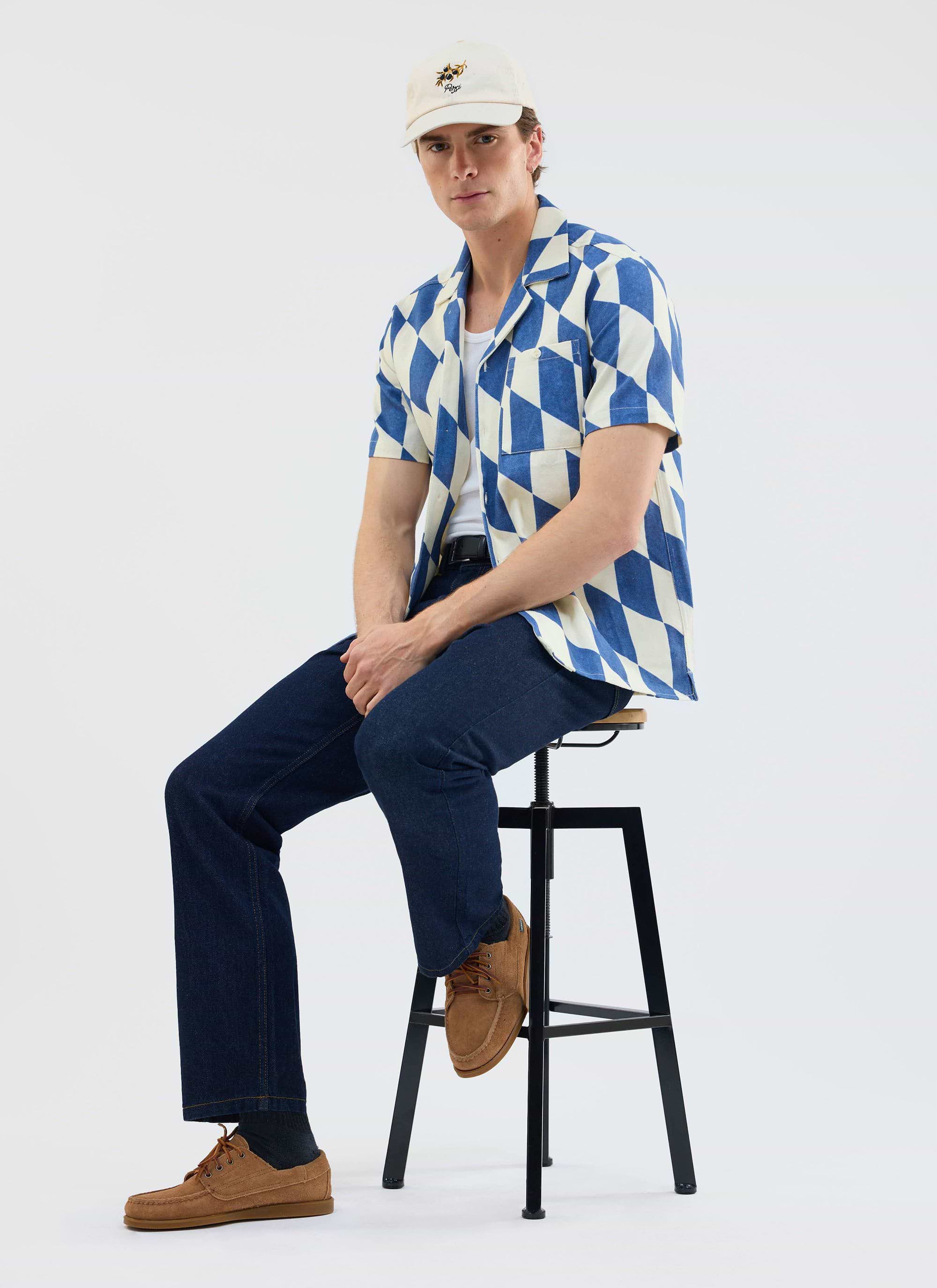 A model wears the Percival Market Diamond Cuban shirt in ecru and navy with dark jeans and a cap while sitting on a stool.