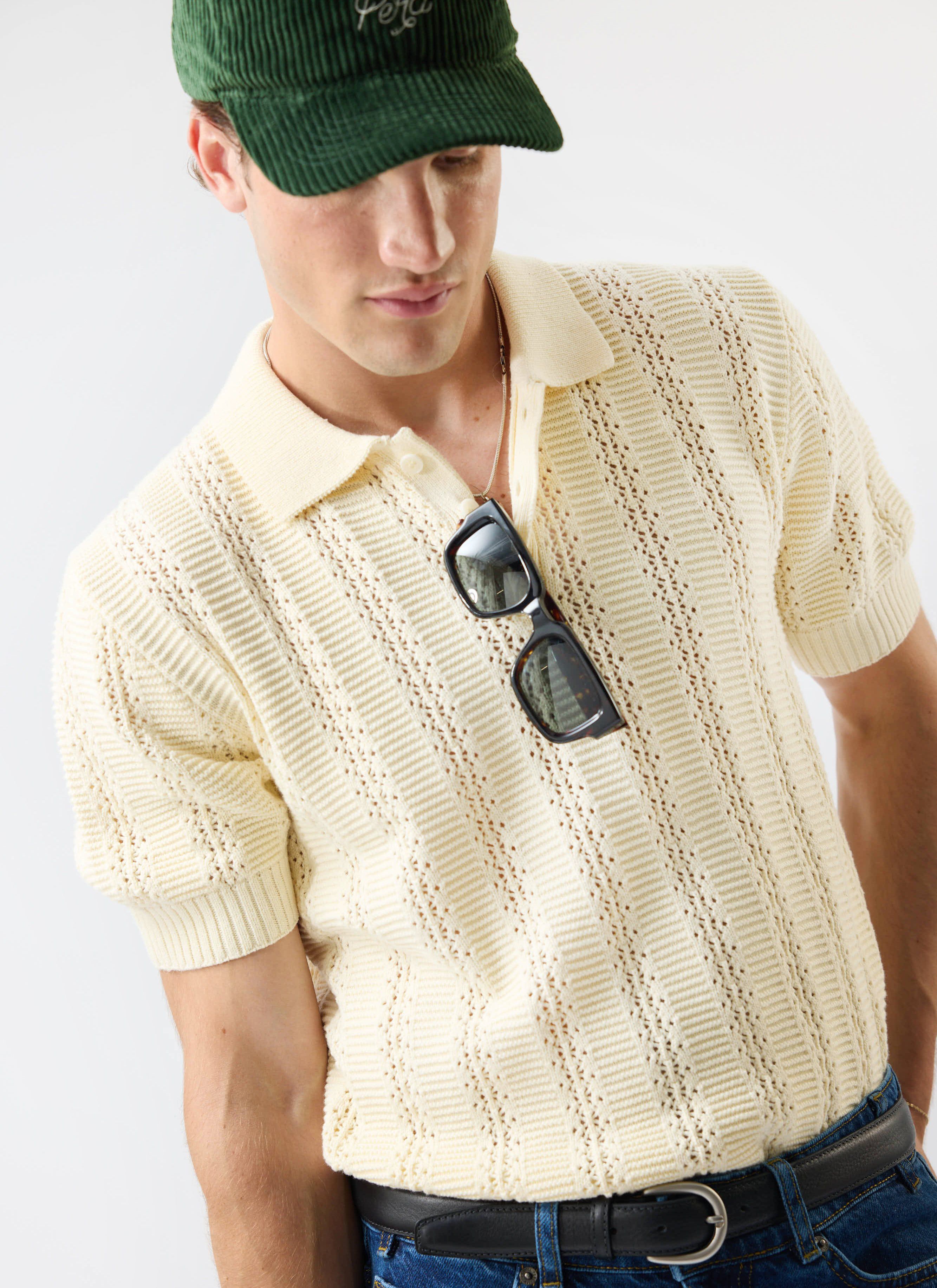 A model wears Percival's ecru cream Addendum Knitted Polo Shirt with a green cap and sunglasses tucked into the placket.