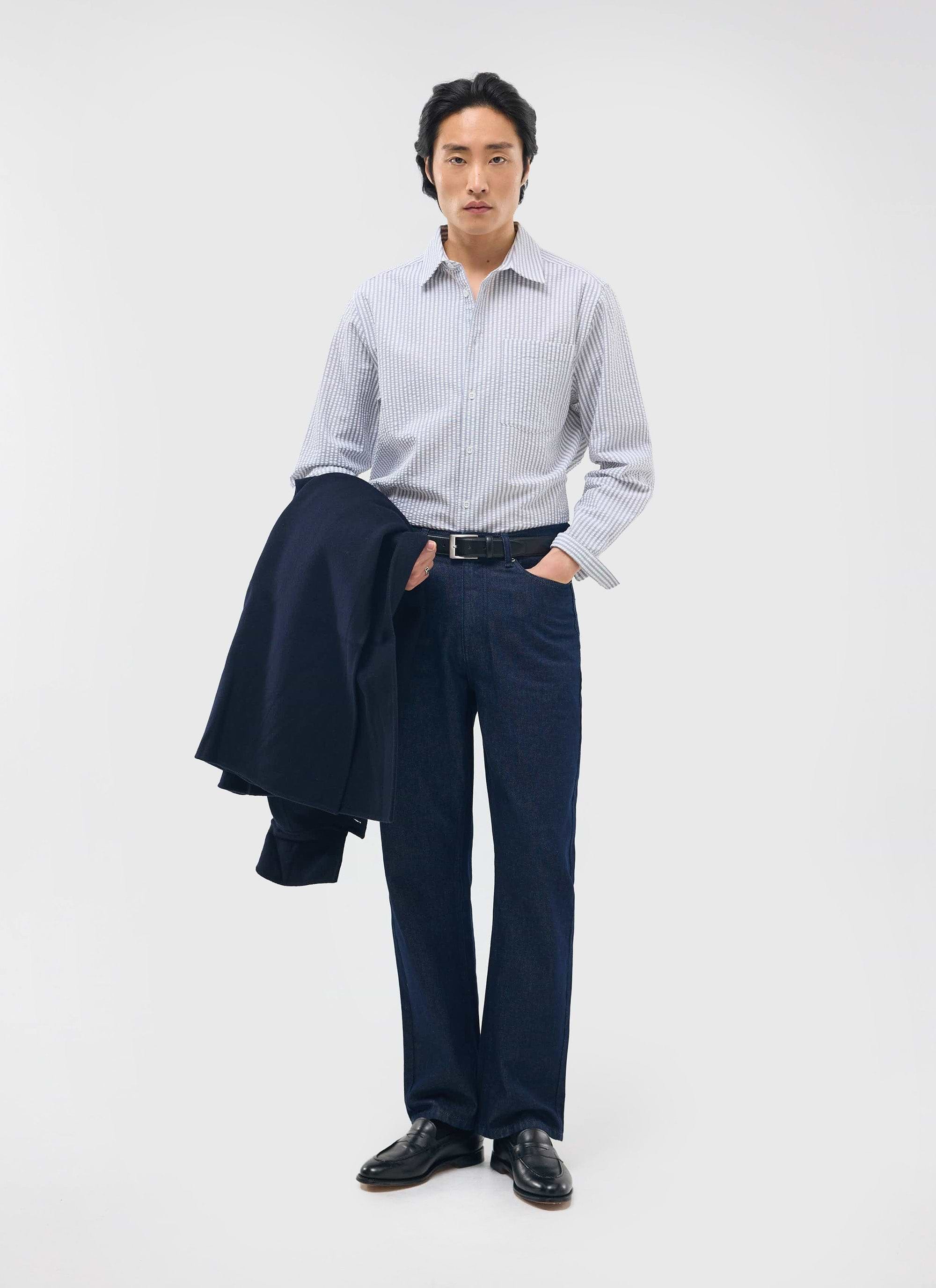 A model in a studio setting wears the Percival Seersucker Stripe Pearce Shirt in grey, with dark jeans and black loafers.