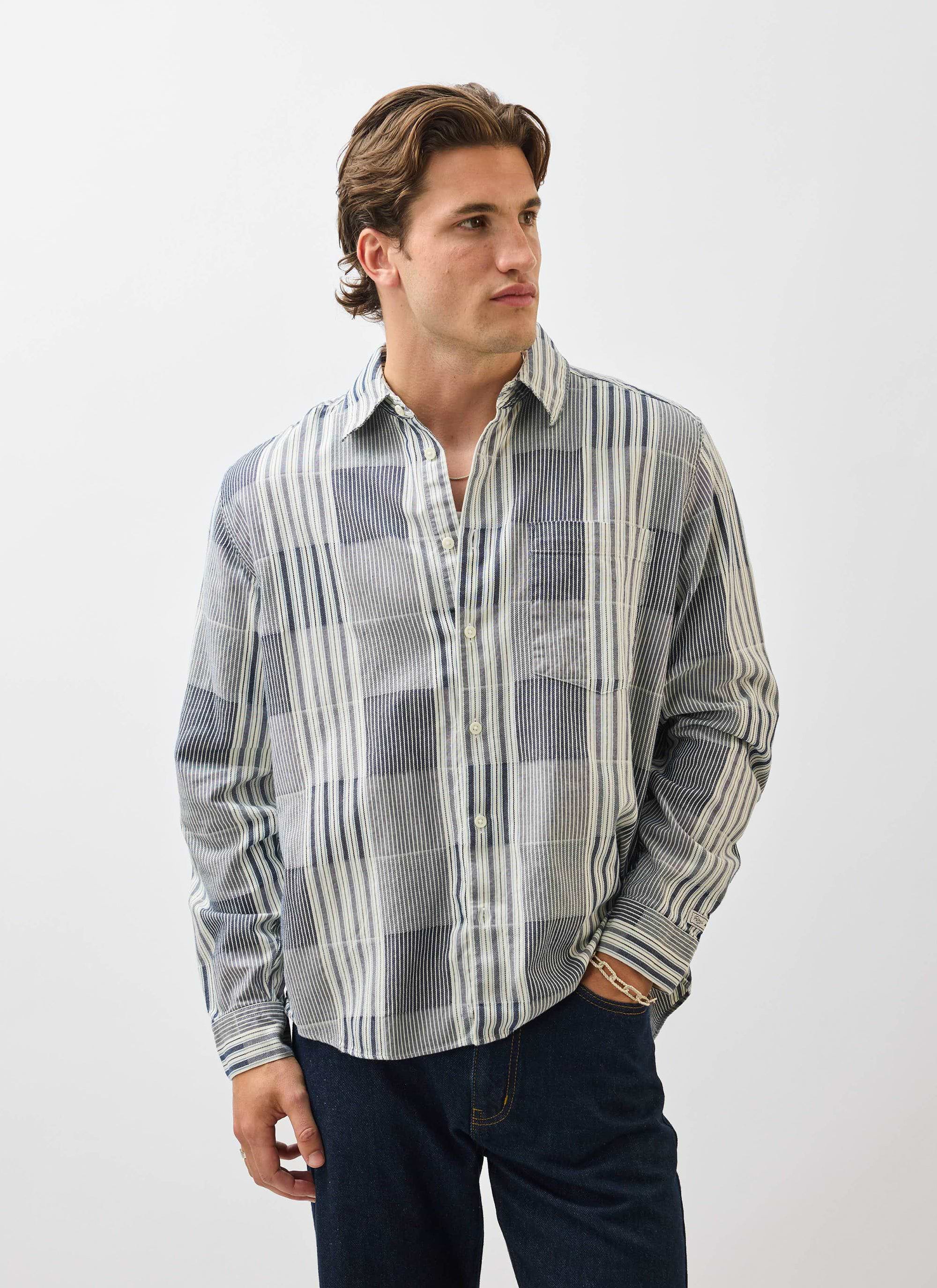 A man models the Percival Epigraph Patchwork Pearce Shirt in blue, worn with dark jeans against a white background.