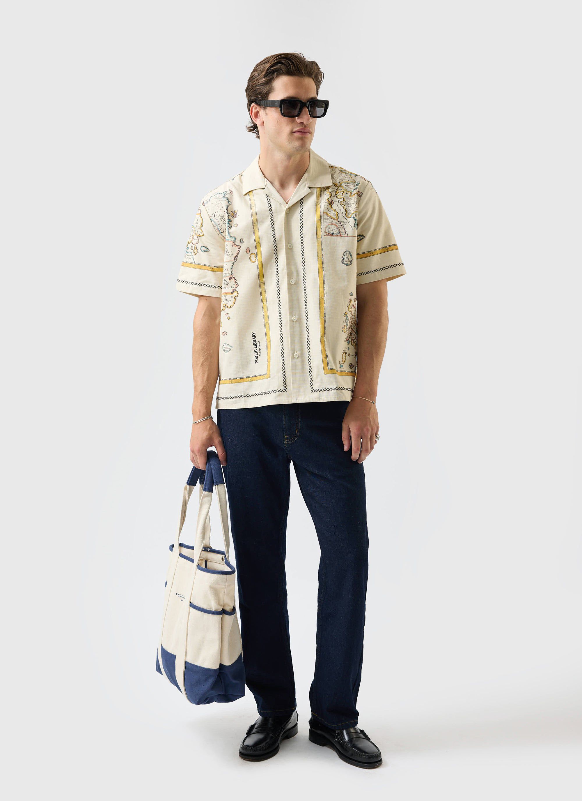 A model wears the Percival Ex Libris Cuban Shirt in ecru, styled with dark jeans, sunglasses, and holding a tote bag.