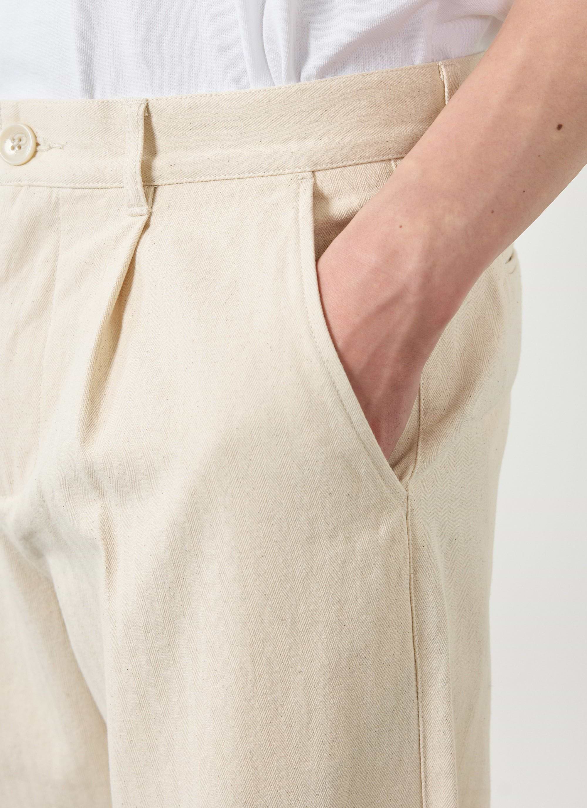 Close up shot of a model wearing the Percival Program Pleated Trousers in ecru cotton, with their hand in the pocket.