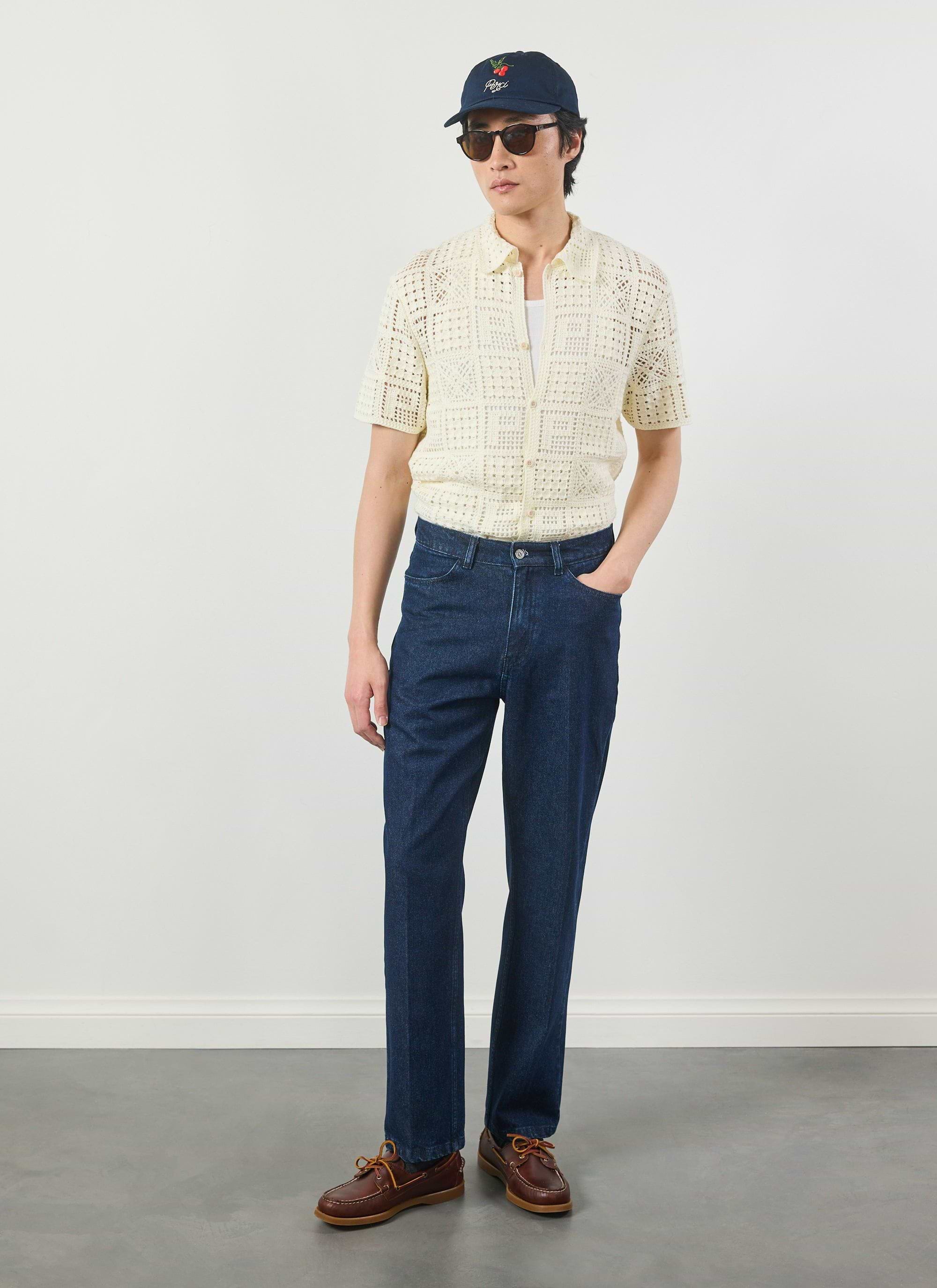 A model wears the Percival Crochet Groundsman Shirt in ecru, styled with dark denim jeans, a navy cap and sunglasses.
